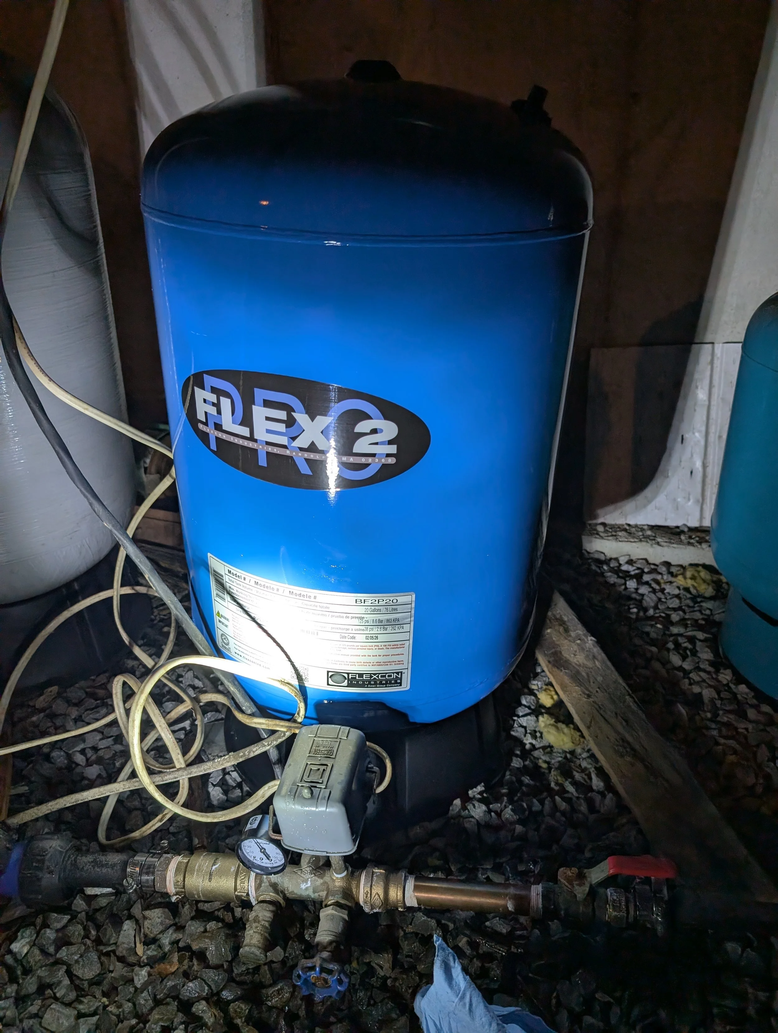 Blue Flexcon water pressure tank connected to plumbing with pipes, valves, and a pressure gauge, situated on gravel. Cables and a power strip are nearby.