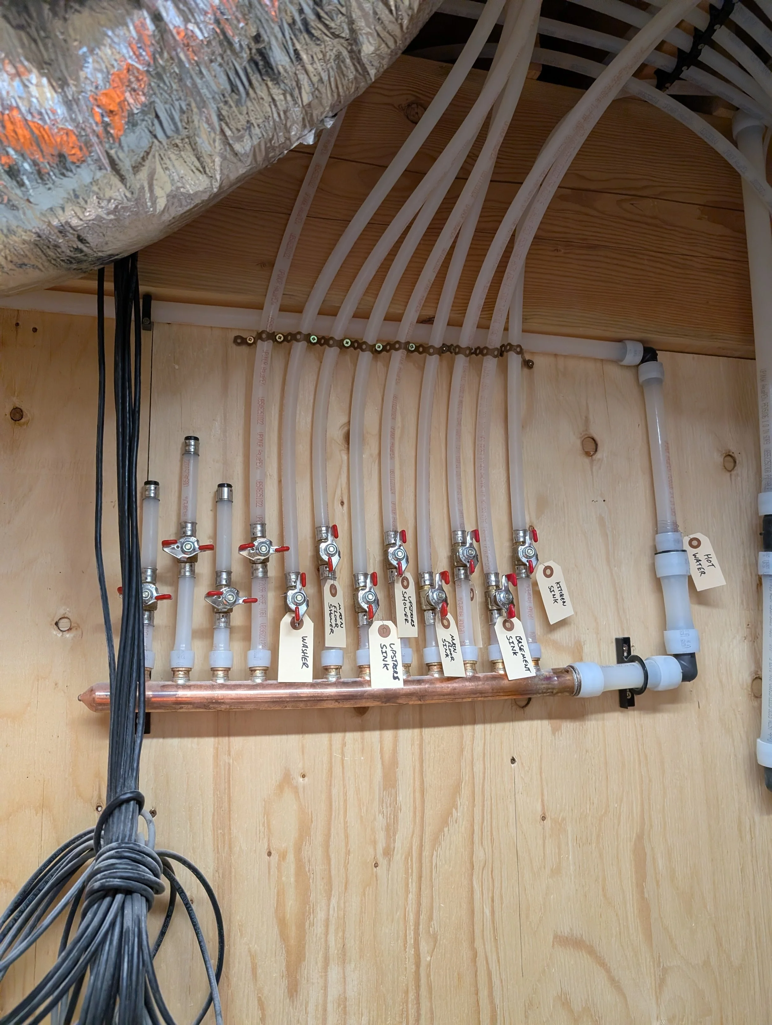 A plumbing system with multiple white pipes labeled for different water lines, a copper manifold, and shut-off valves, installed against a wooden wall in a building or utility room.