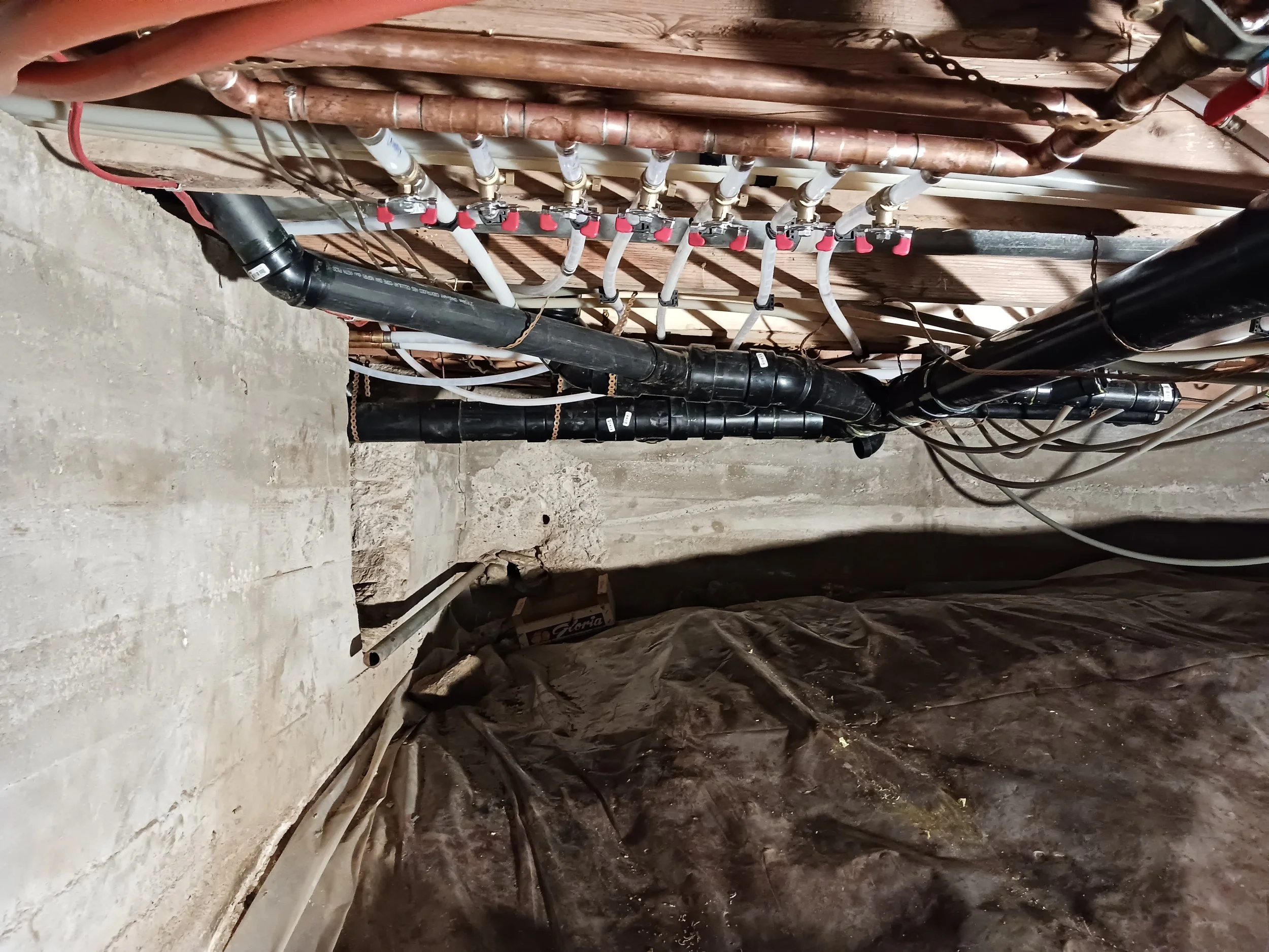 A plumbing installation in a basement or crawl space, showing copper pipes, black PVC pipes, and white plastic pipes connected with various fittings, with a concrete floor and exposed wooden ceiling.