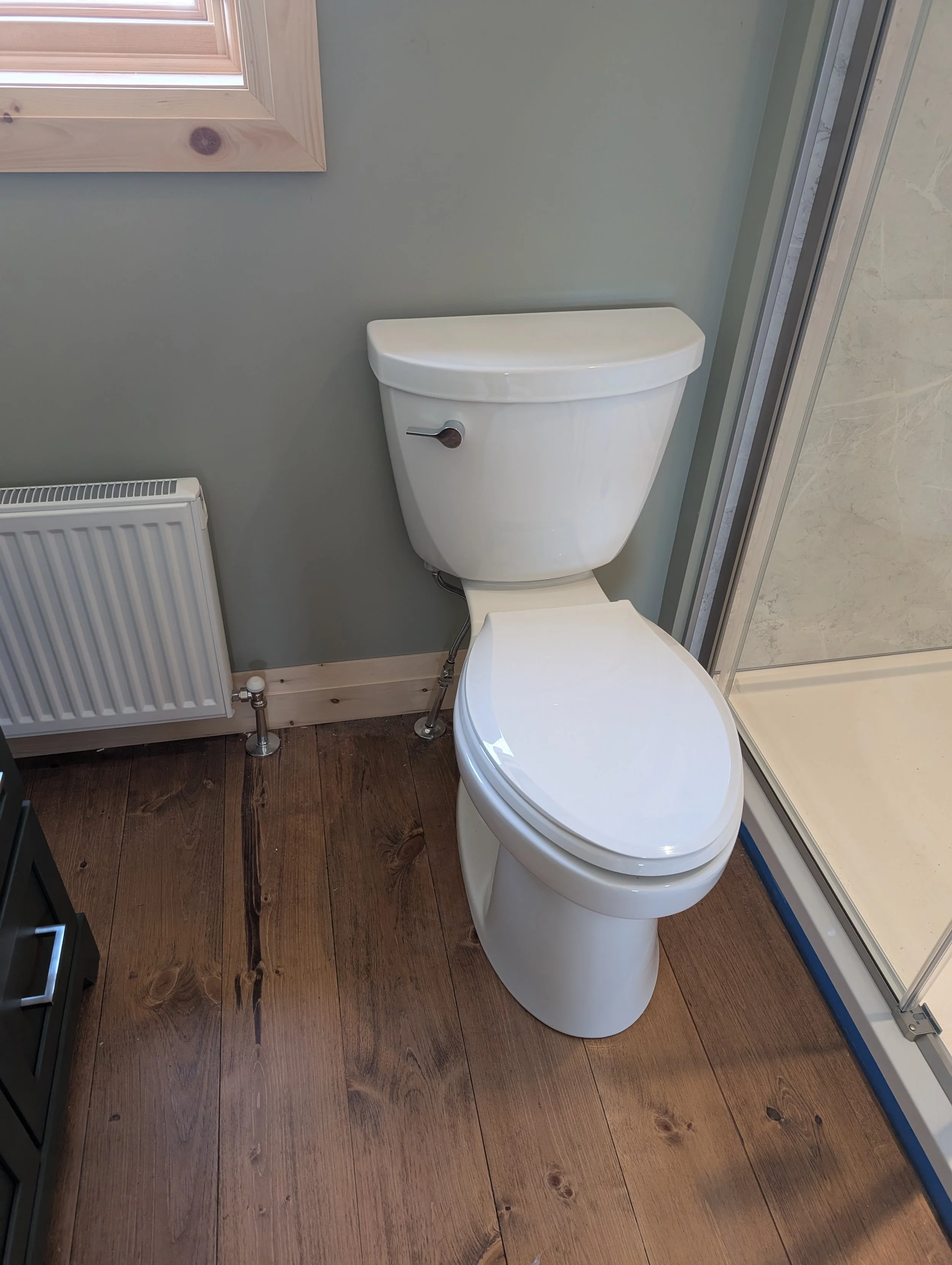 Bathroom with a white toilet, a window, radiator, and a shower enclosure on a wooden floor.