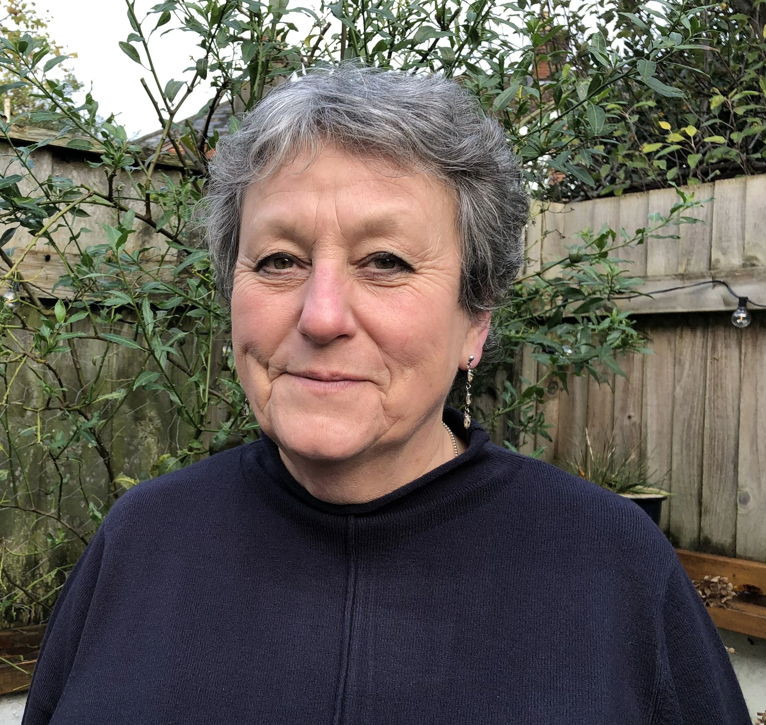 Headshot of Caroline smiling at the camera, with short grey hair and wearing a navy blue jumper, standing in front of a tree.