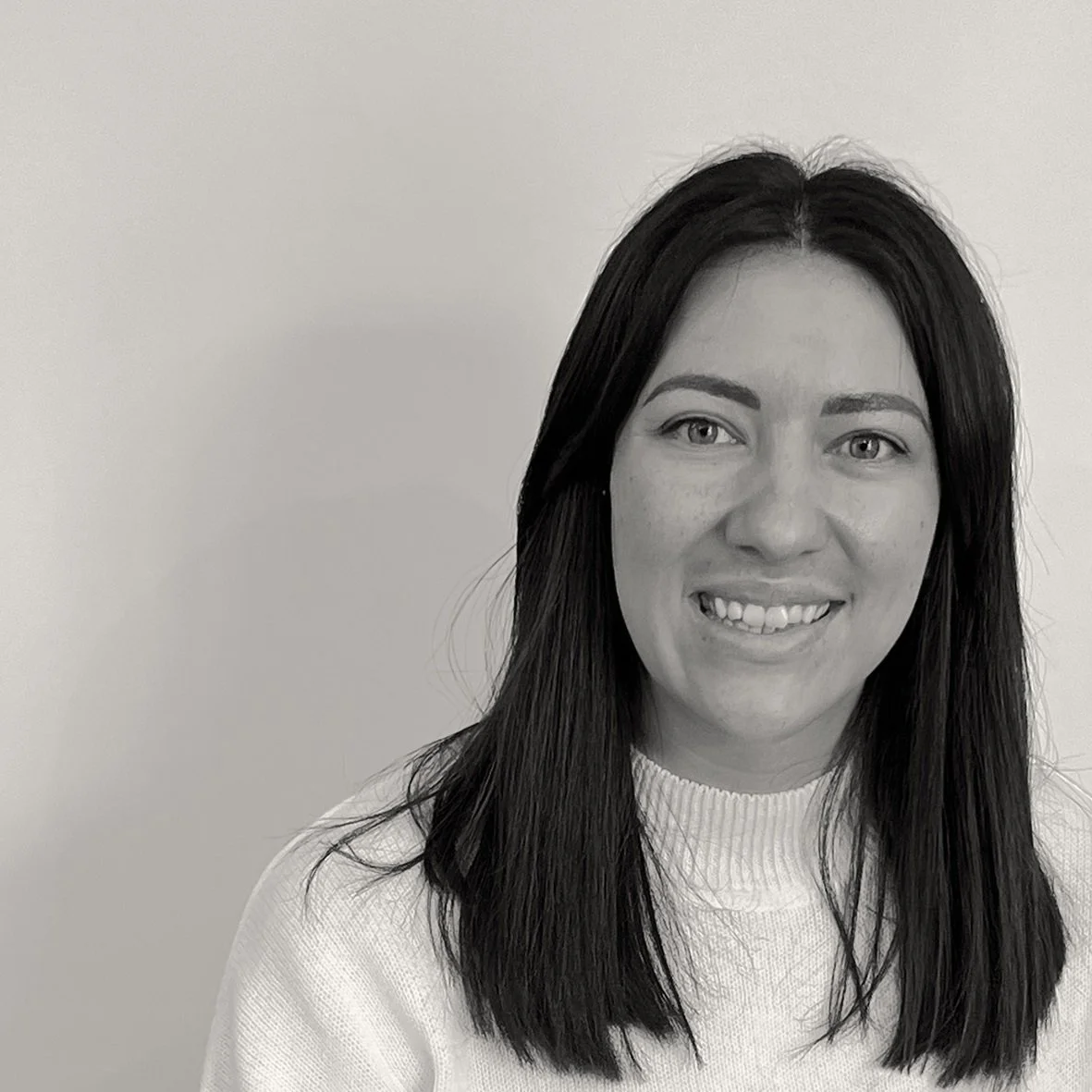 Black and white headshot of Poppy, with dark hair, smiling while looking at the camera.