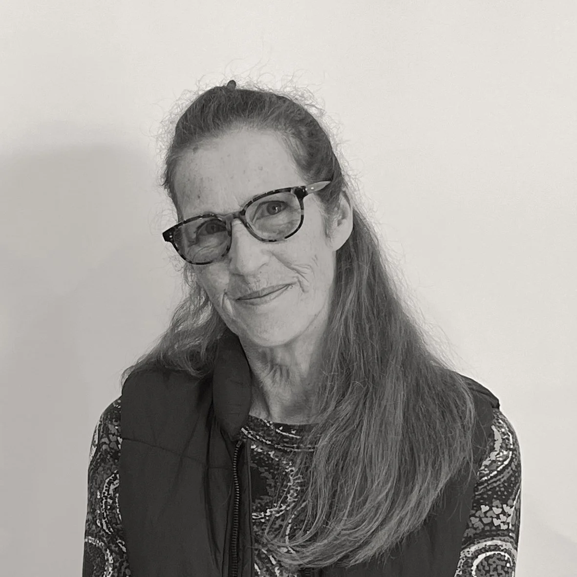 Black and white headshot of Polly, with half-up, half-down hair draped over one shoulder, smiling and wearing glasses.