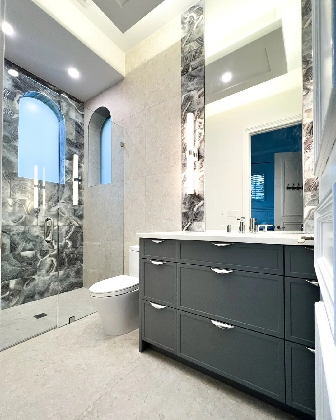 Modern bathroom with a walk-in shower featuring marble tiles, a gray vanity with six drawers, a large mirror, and a toilet beside the vanity.