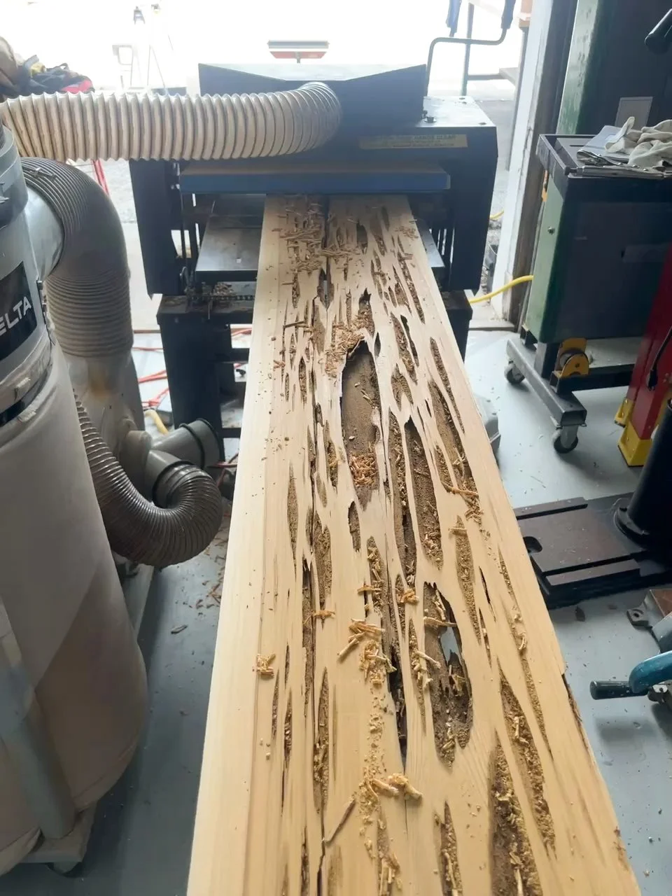 A woodworking machine in a workshop actively carving a large wooden beam, creating intricate and elongated patterns. Sawdust and wood shavings are scattered on the beam and around the machine.