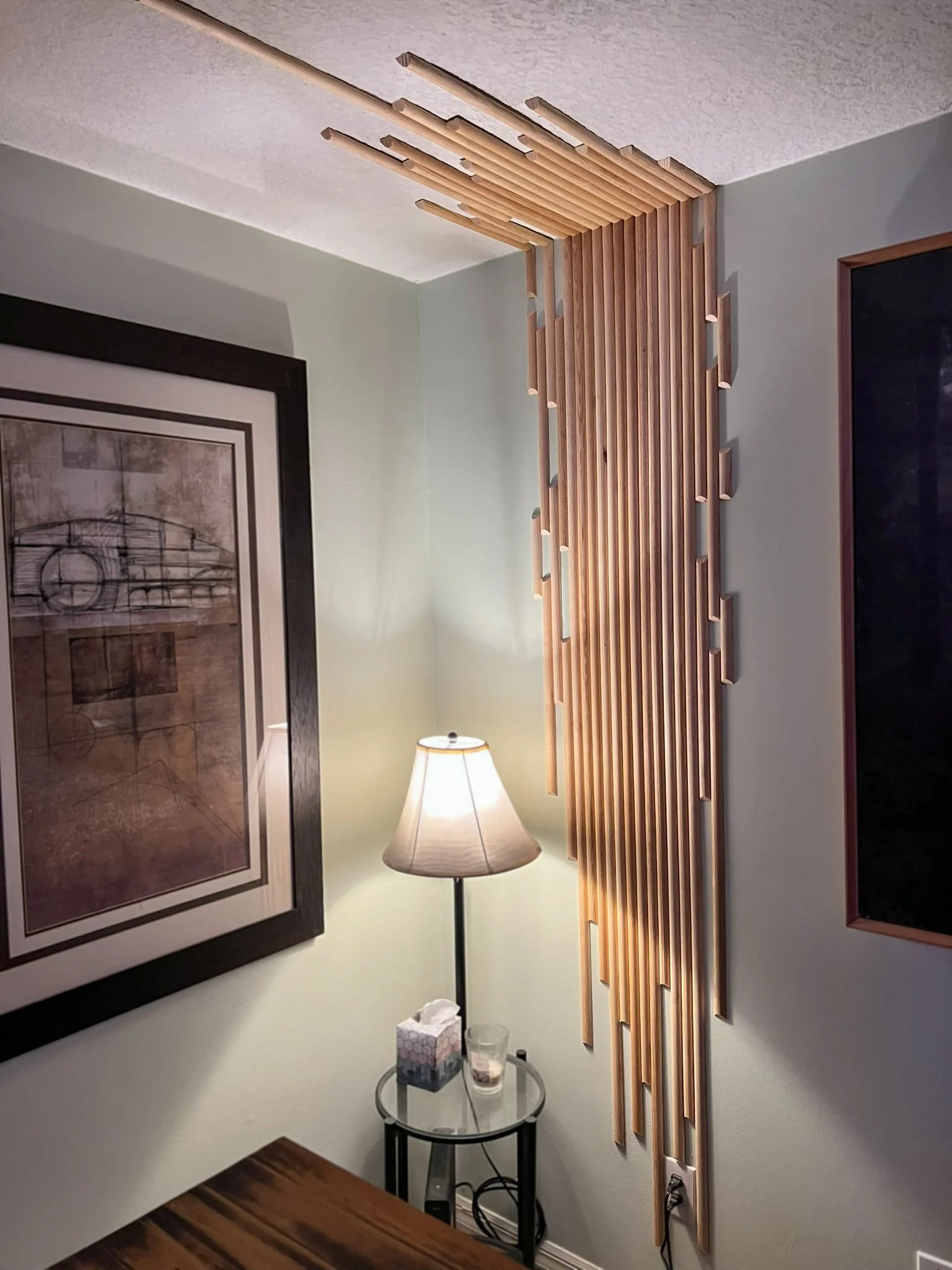 Wooden wall art installation with vertical and horizontal slats in a corner of a room, illuminated by a floor lamp, with a framed picture on the adjacent wall.