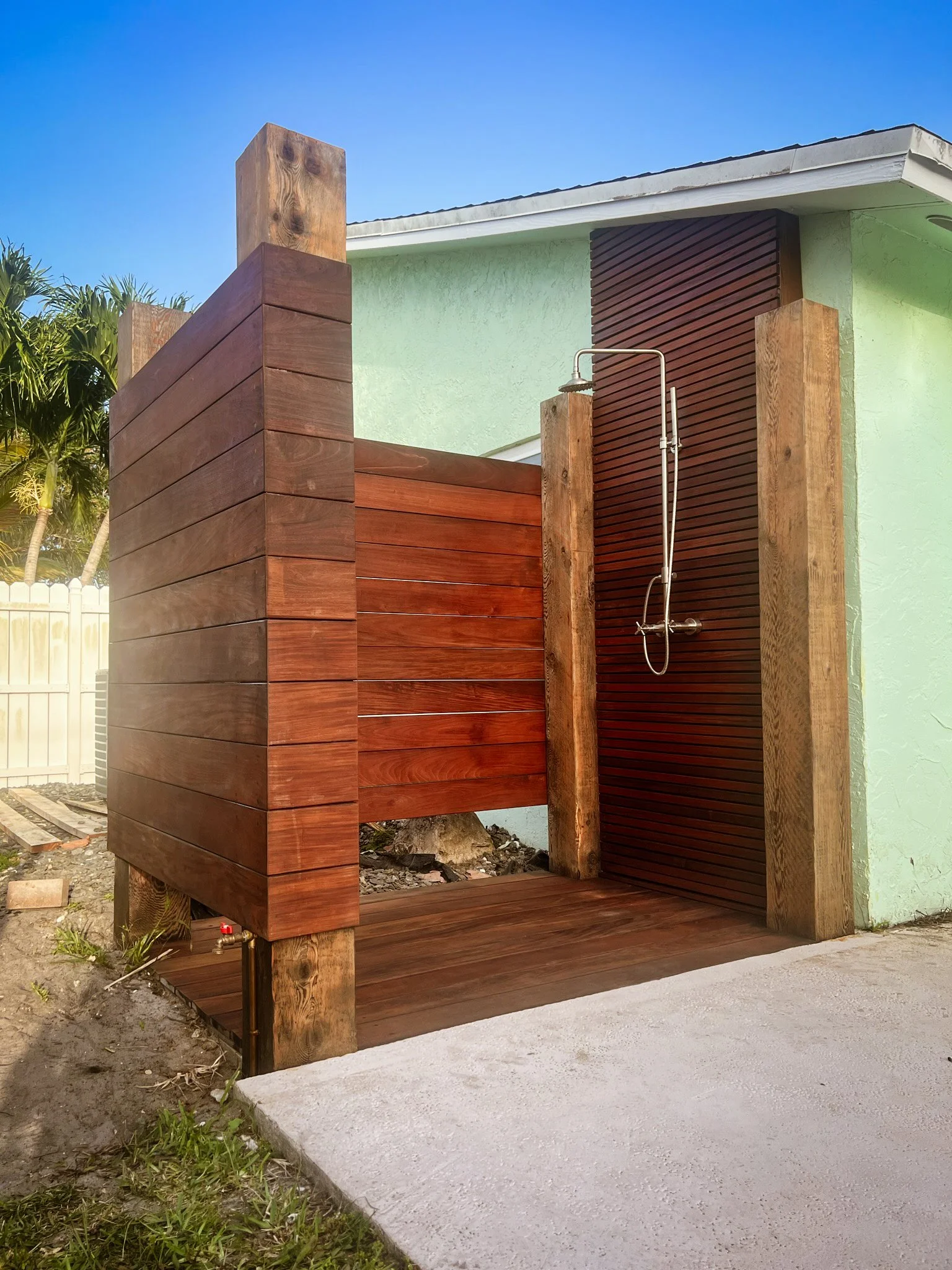 Outdoor wooden shower with a metal showerhead, situated next to a green house wall, under a clear blue sky.