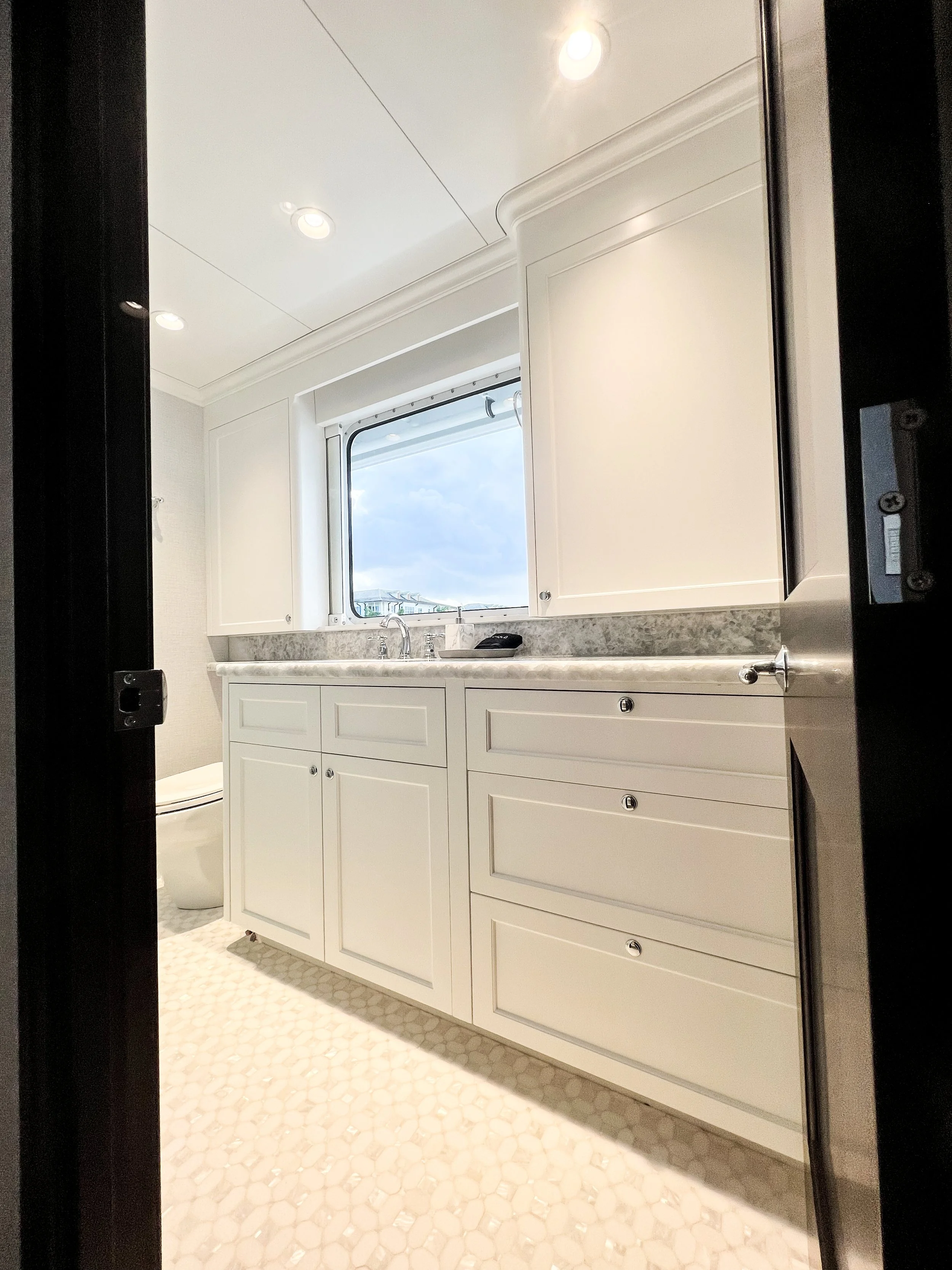 A bright bathroom with white cabinets under a marble countertop, above which a window shows a partly cloudy sky. Recessed lighting in the ceiling illuminates the space, and a toilet is partially visible on the left side.