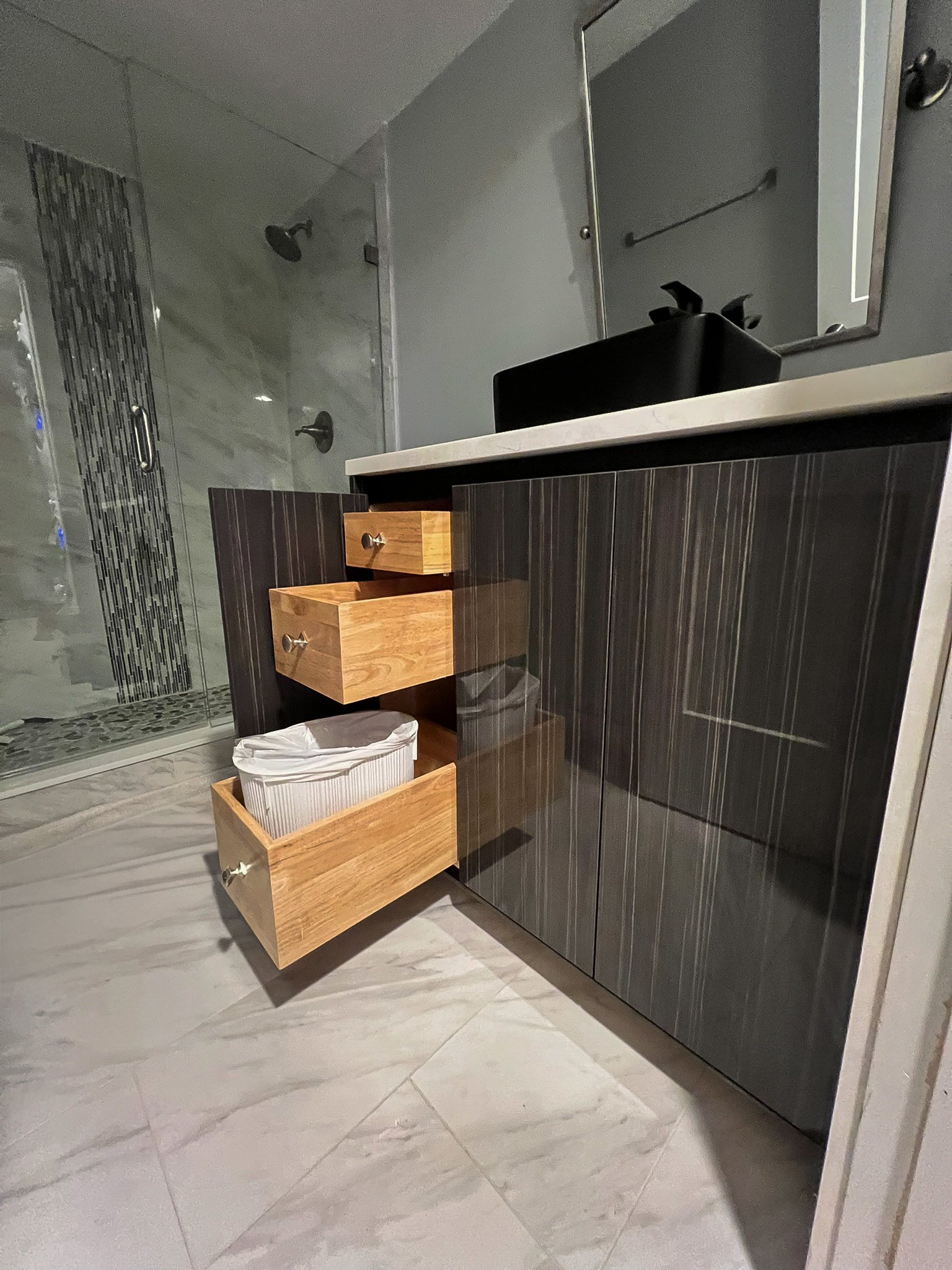 Bathroom vanity with open drawers, a trash can, and a mirror above a black sink.