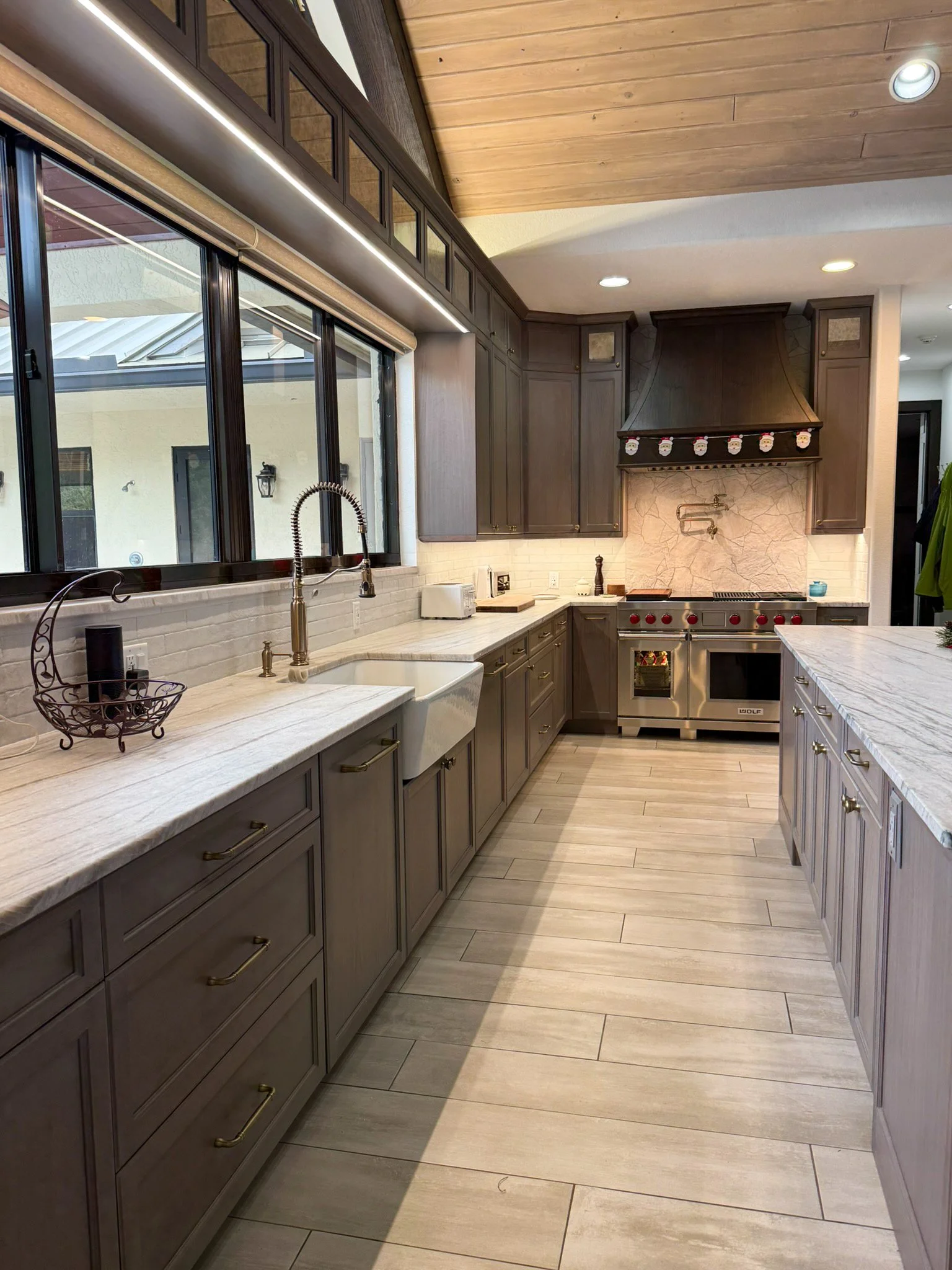 Kitchen with dark wood cabinets, white marble countertops, a large window, stainless steel stove, and a farmhouse sink.