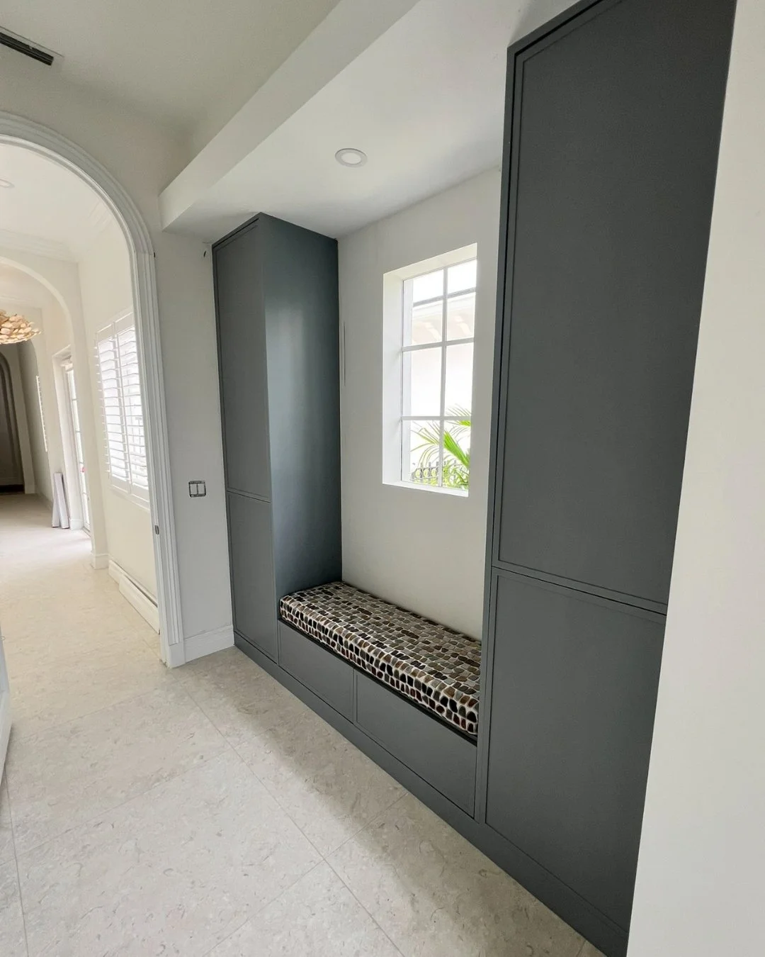 Modern built-in gray cabinetry with a cushioned bench seat in a bright hallway with a window, white walls, and tiled floor.