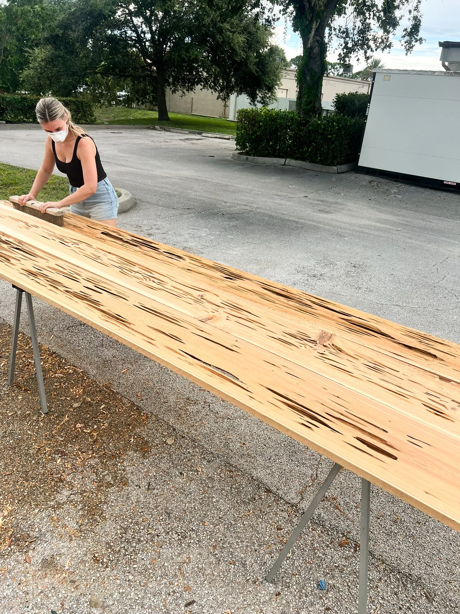 A woman wearing a face mask and black tank top stands outside at a wooden workbench, working on a large piece of wood with a sander. The background includes trees, bushes, and a parking lot.