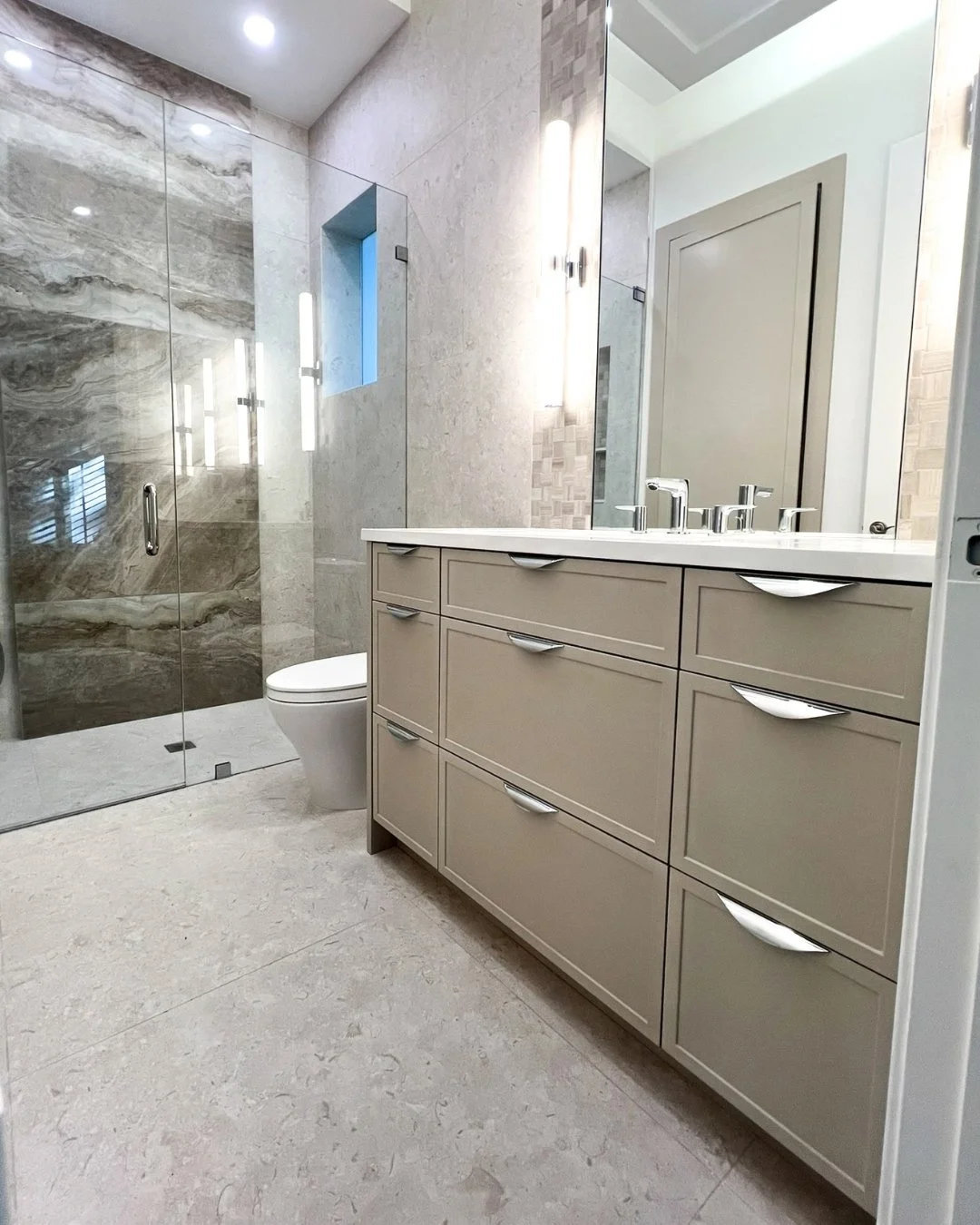 Modern bathroom with beige cabinetry, large mirror, and a glass-enclosed shower with marble walls and a small window.