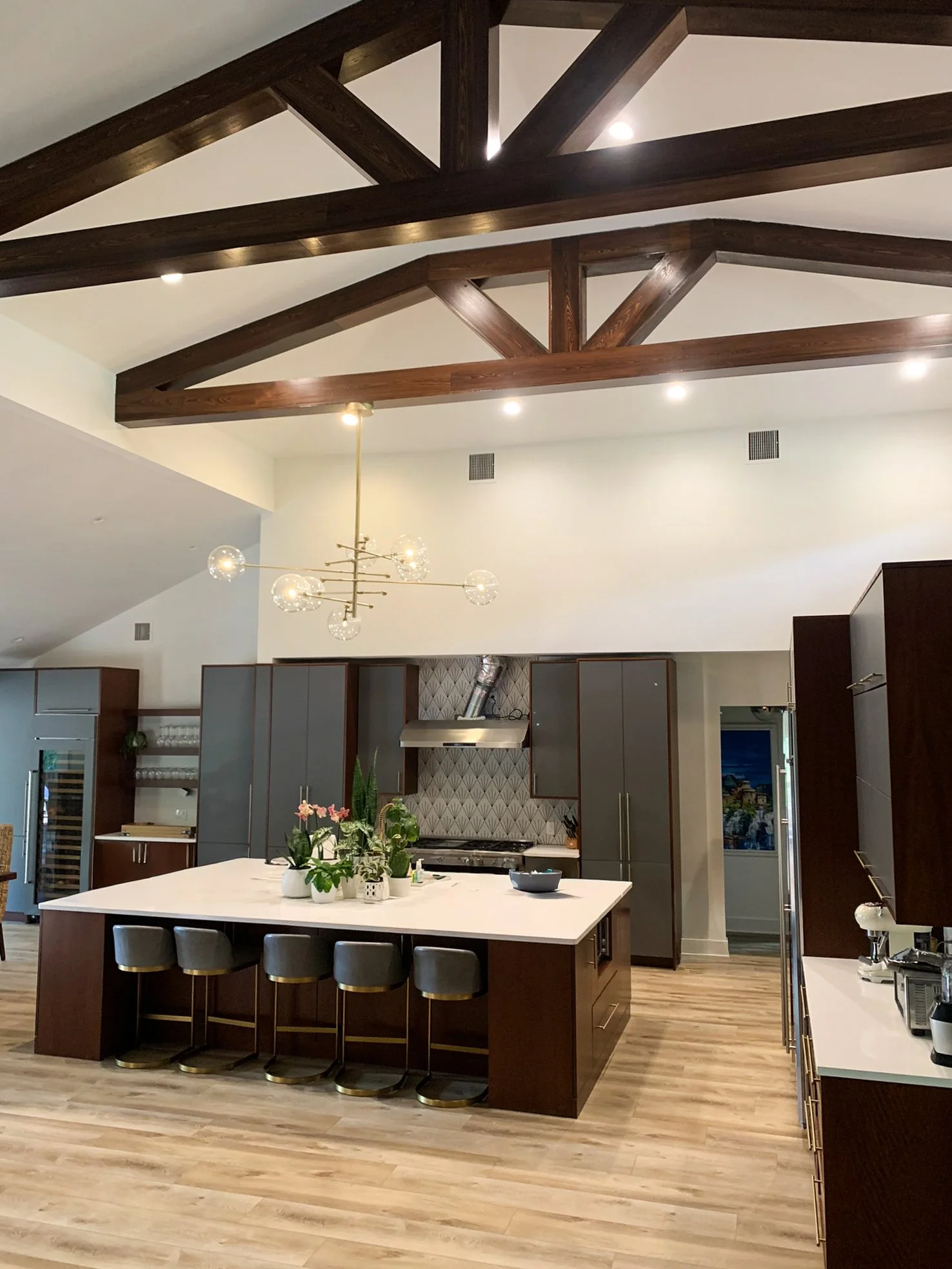 Modern kitchen with dark wood cabinets, a large white island with seating, a gold chandelier, and exposed wooden ceiling beams.