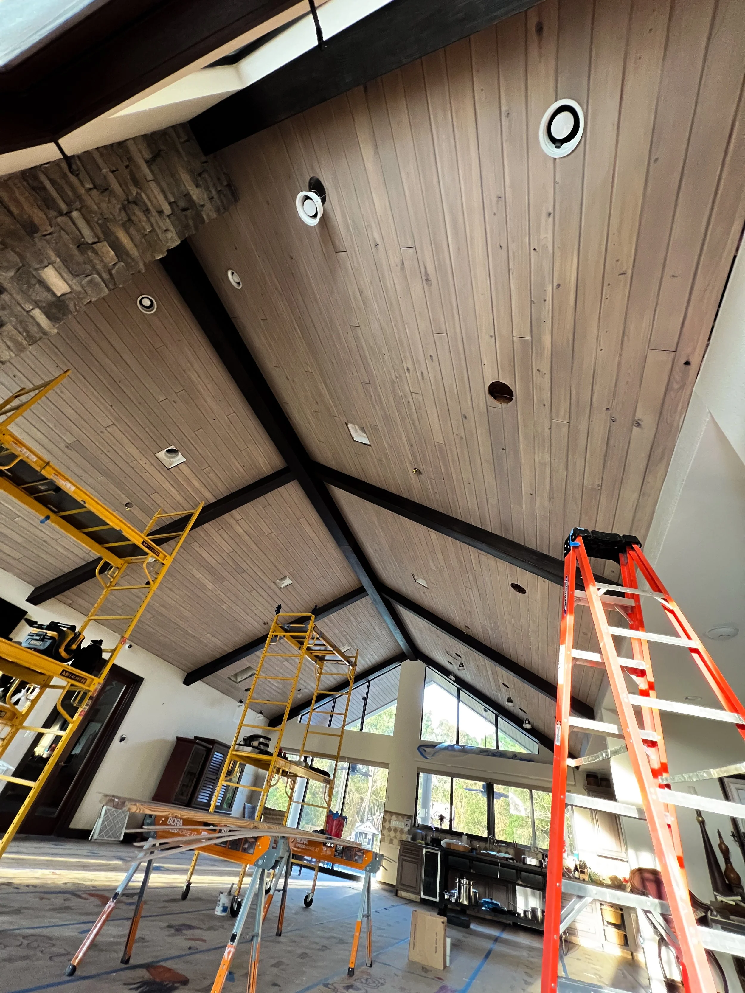 Interior view of a house under construction with a vaulted wooden ceiling, scaffolding, a ladder, and construction tools.
