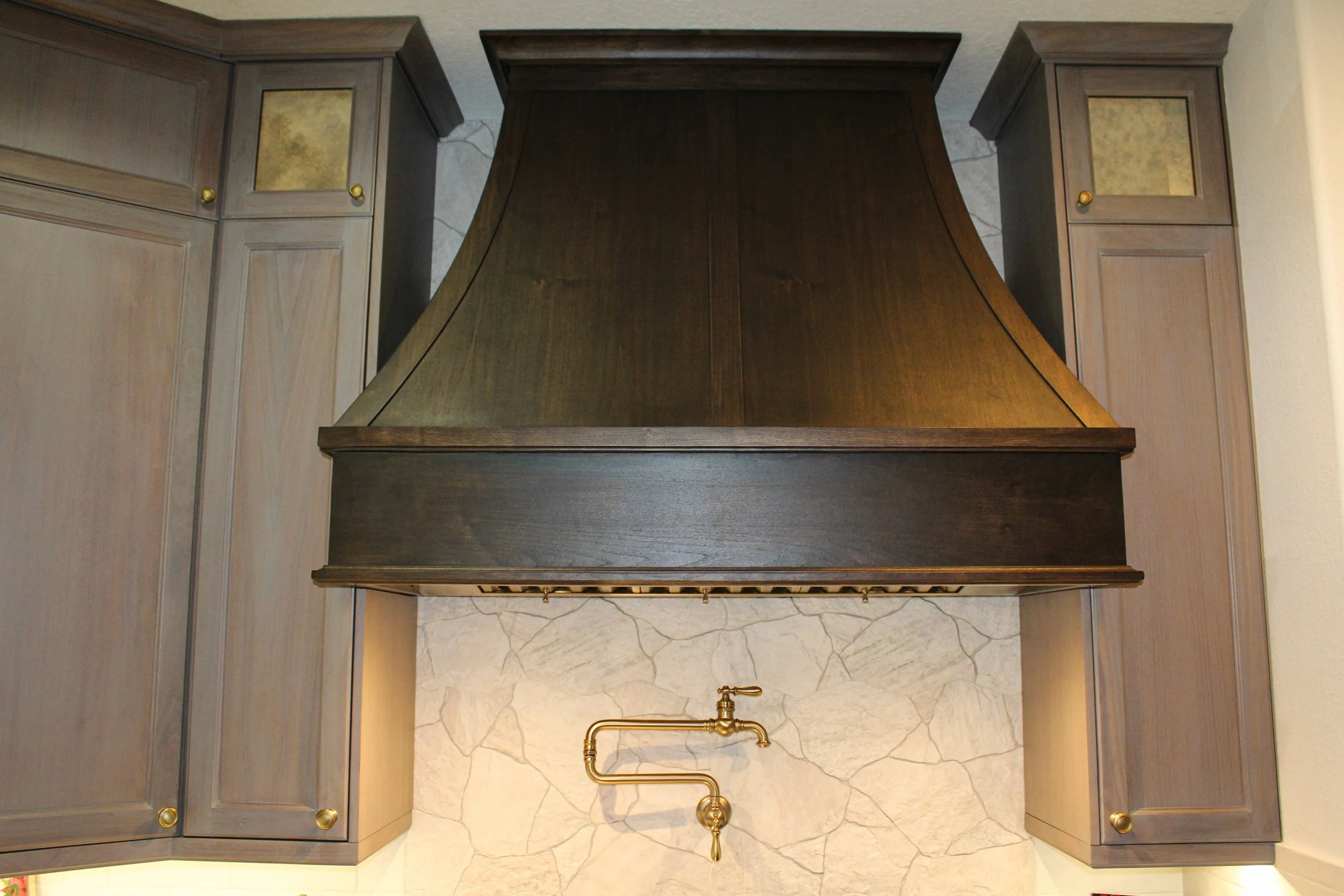 Wood kitchen range hood above a stone backsplash with a brass faucet and pipe.