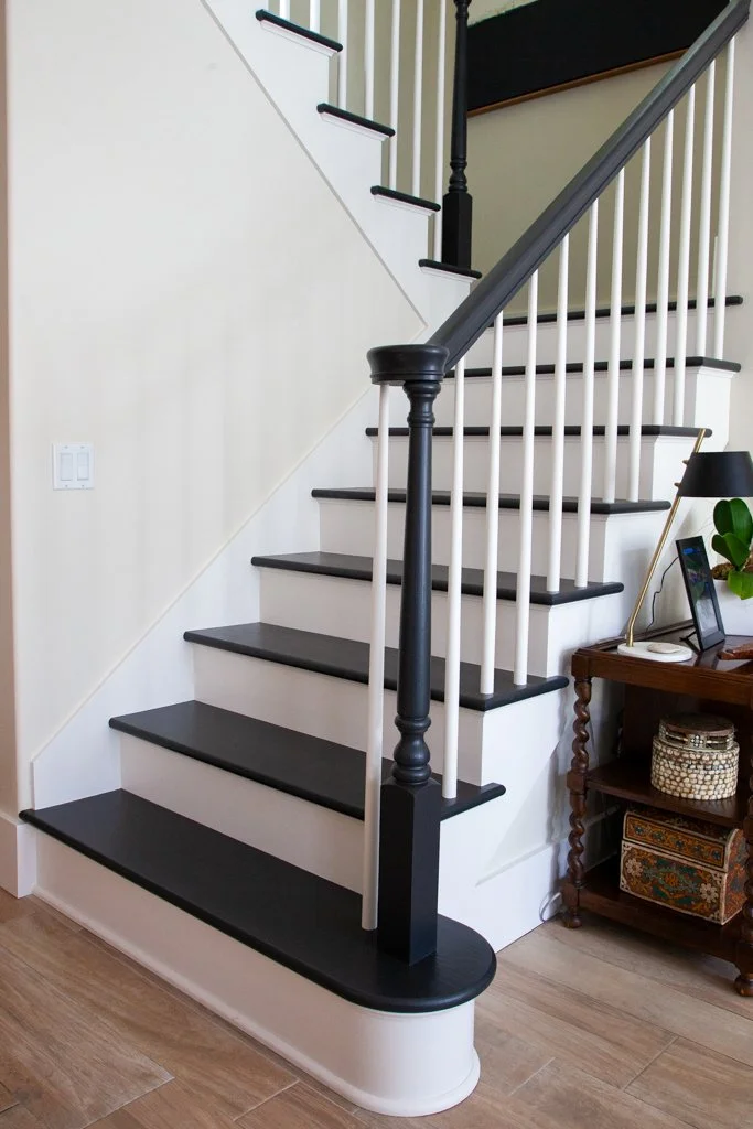 A staircase with black and white colors, black handrail, and decorative newel post, next to a small wooden side table with items on top and a storage basket underneath.