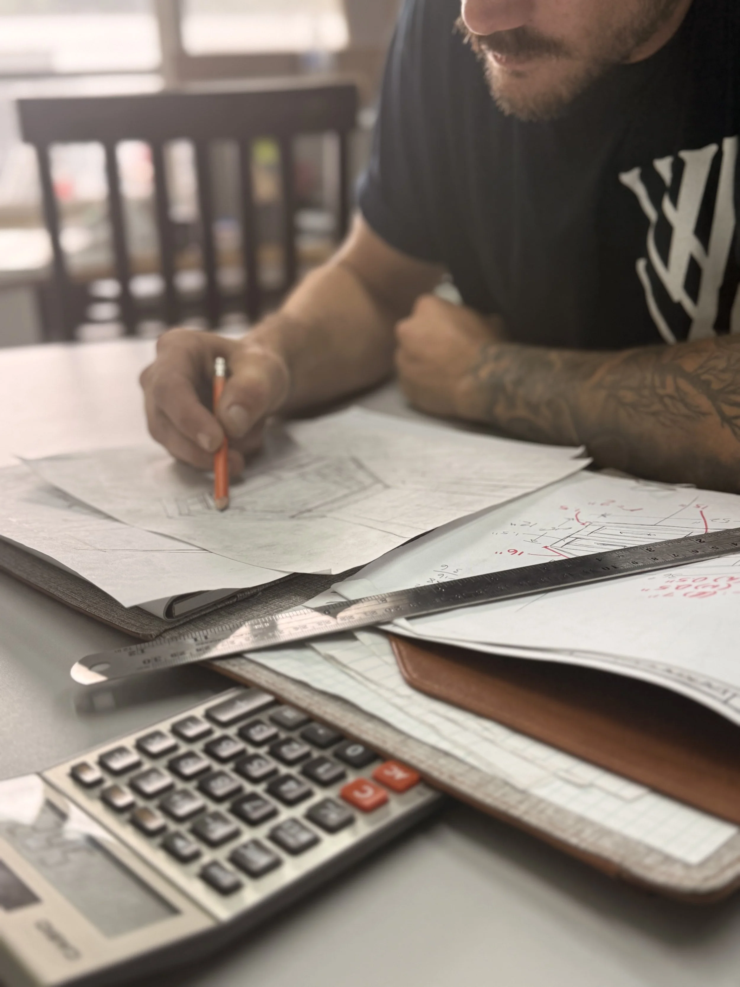 A person at a desk reviewing architectural plans with a pencil, surrounded by papers, a calculator, and a ruler.