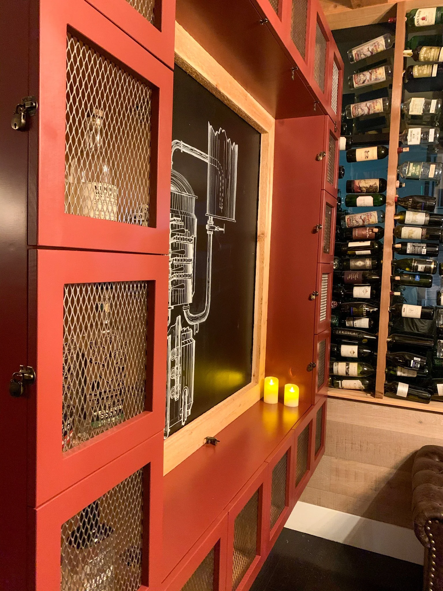 A wine cellar with red cabinets, a chalkboard with a wine barreling diagram, two lit candles, and a wine rack filled with bottles.