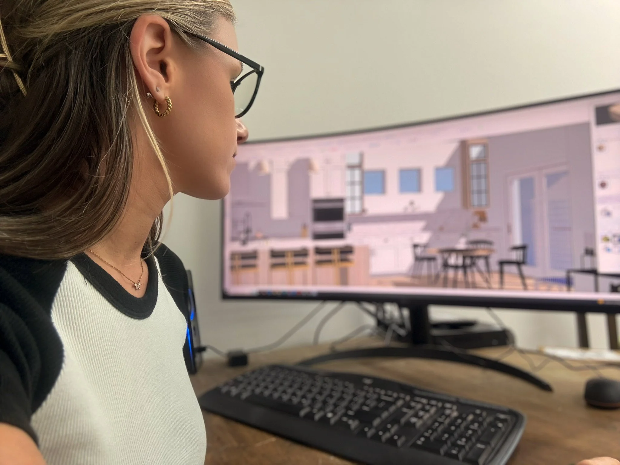 Side profile of a woman with blonde hair, wearing glasses, earrings, and a necklace, working on a computer with architectural plans or designs on the screen.