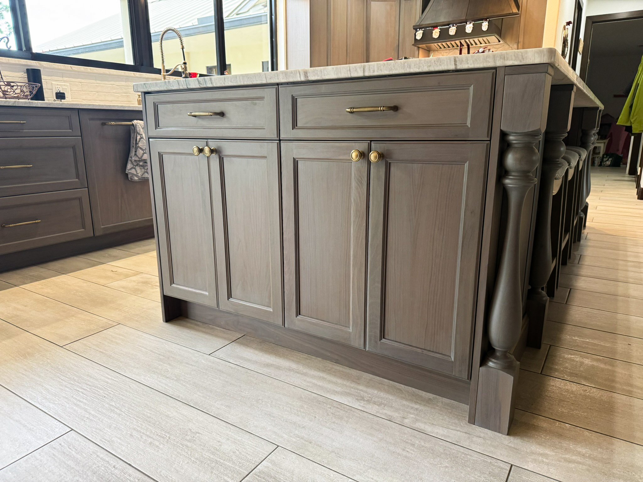 Kitchen island with beige cabinet doors, gold handles, marble countertop, and turned wood legs, with gray cabinetry and a window in the background.