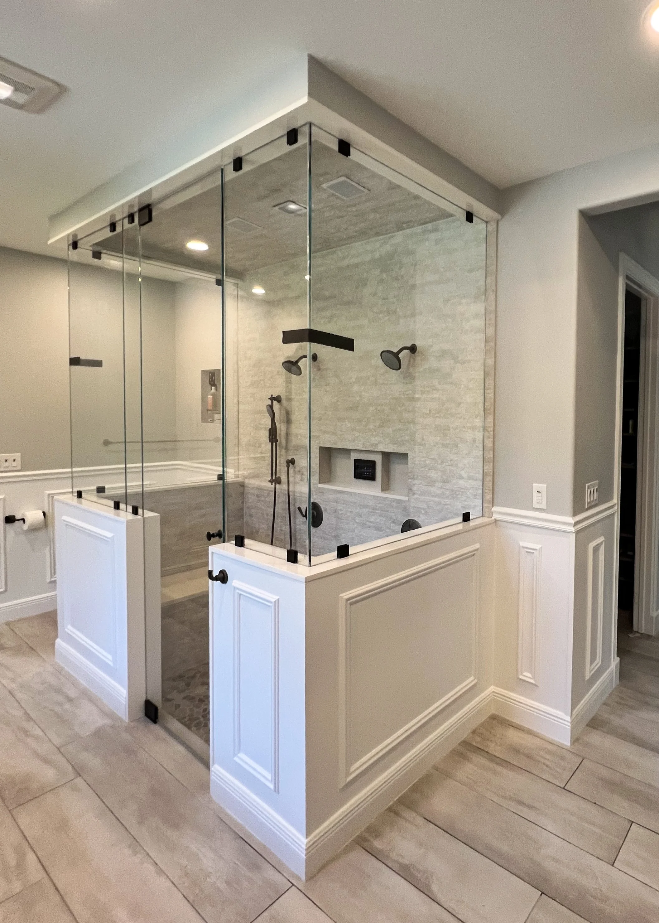 Glass-enclosed shower with multiple showerheads and a built-in niche for toiletries, located in a modern bathroom with light-colored tiled floors and wainscoting on the walls.