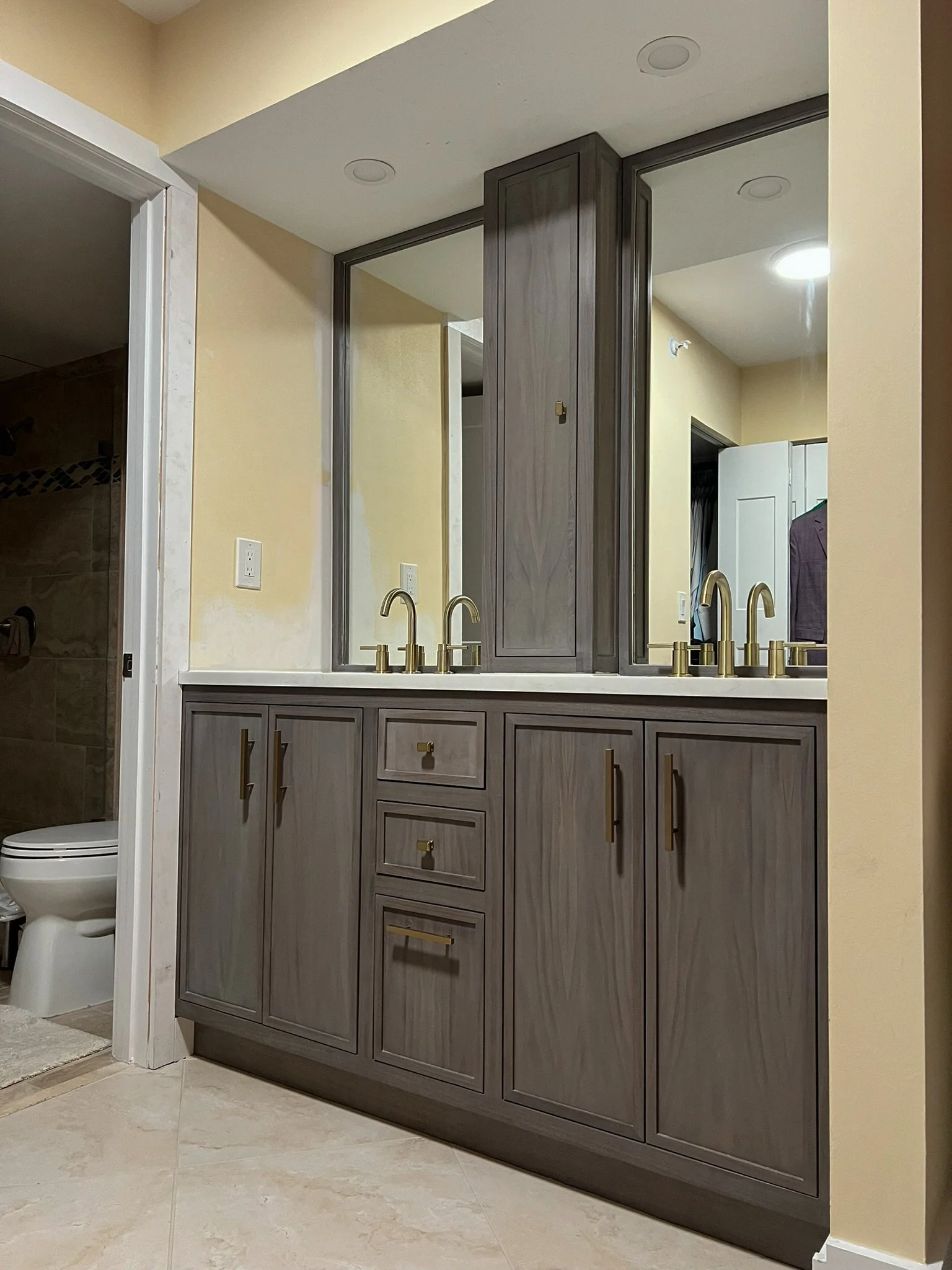 A bathroom vanity with gray wooden cabinets, four drawers, and two sinks with gold faucets. A large mirror spans the length of the vanity, and an adjacent visible bathroom with a toilet and shower.