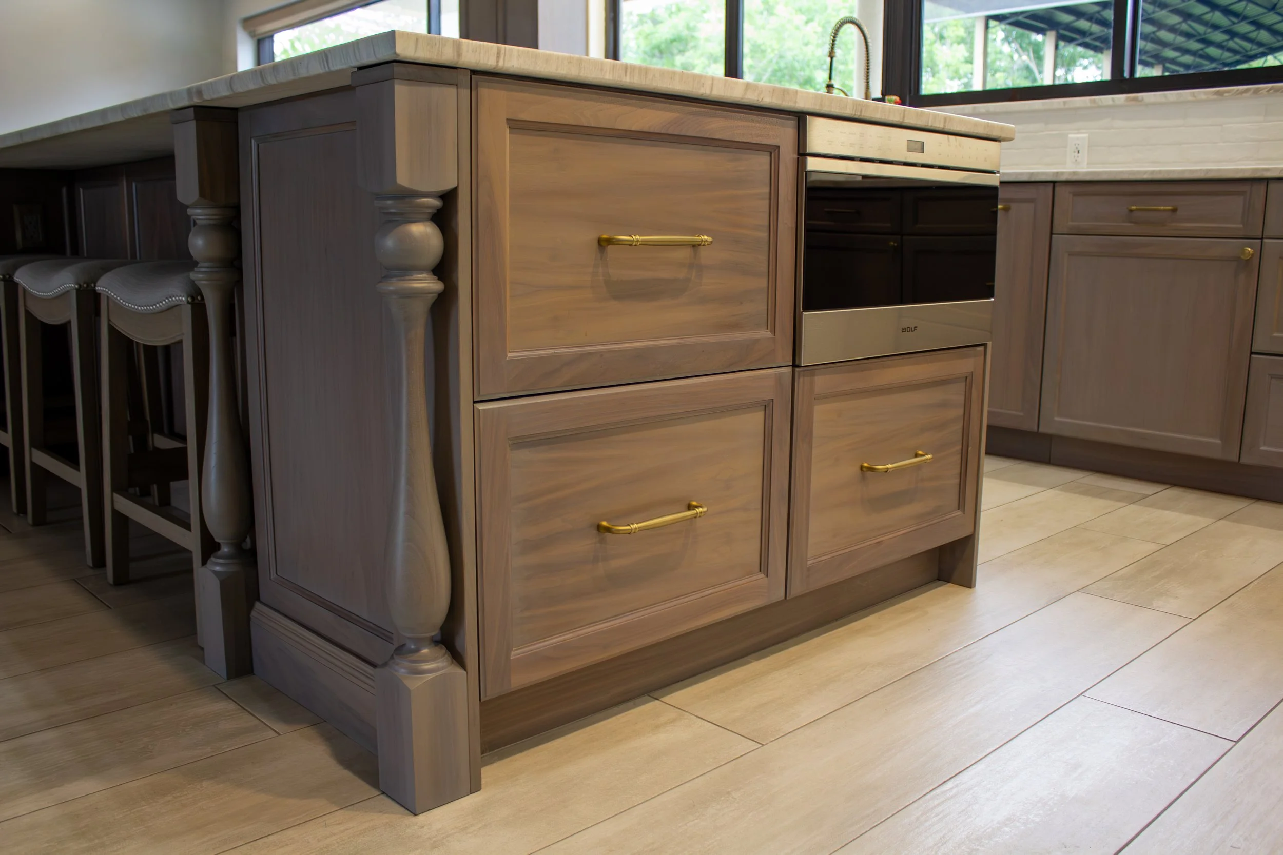 Kitchen island with four drawers featuring gold handles, topped with a beige countertop, with dark and light wood cabinets and windows in the background.