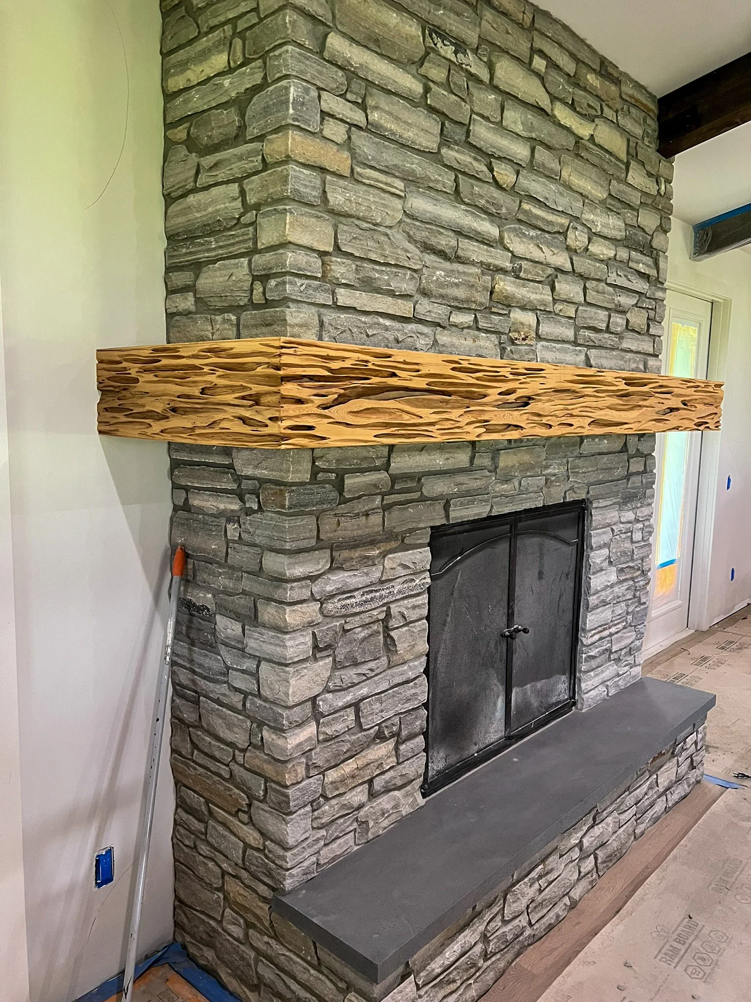 A stone fireplace with a gray stone surround and a wooden mantelpiece in a room under construction, with plastic protective coverings and construction materials visible.