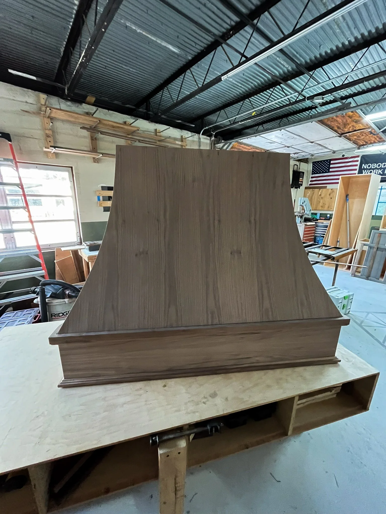 A woodworking workshop with a partially assembled wooden structure on a workbench, surrounded by tools and lumber, with an American flag and a sign in the background.