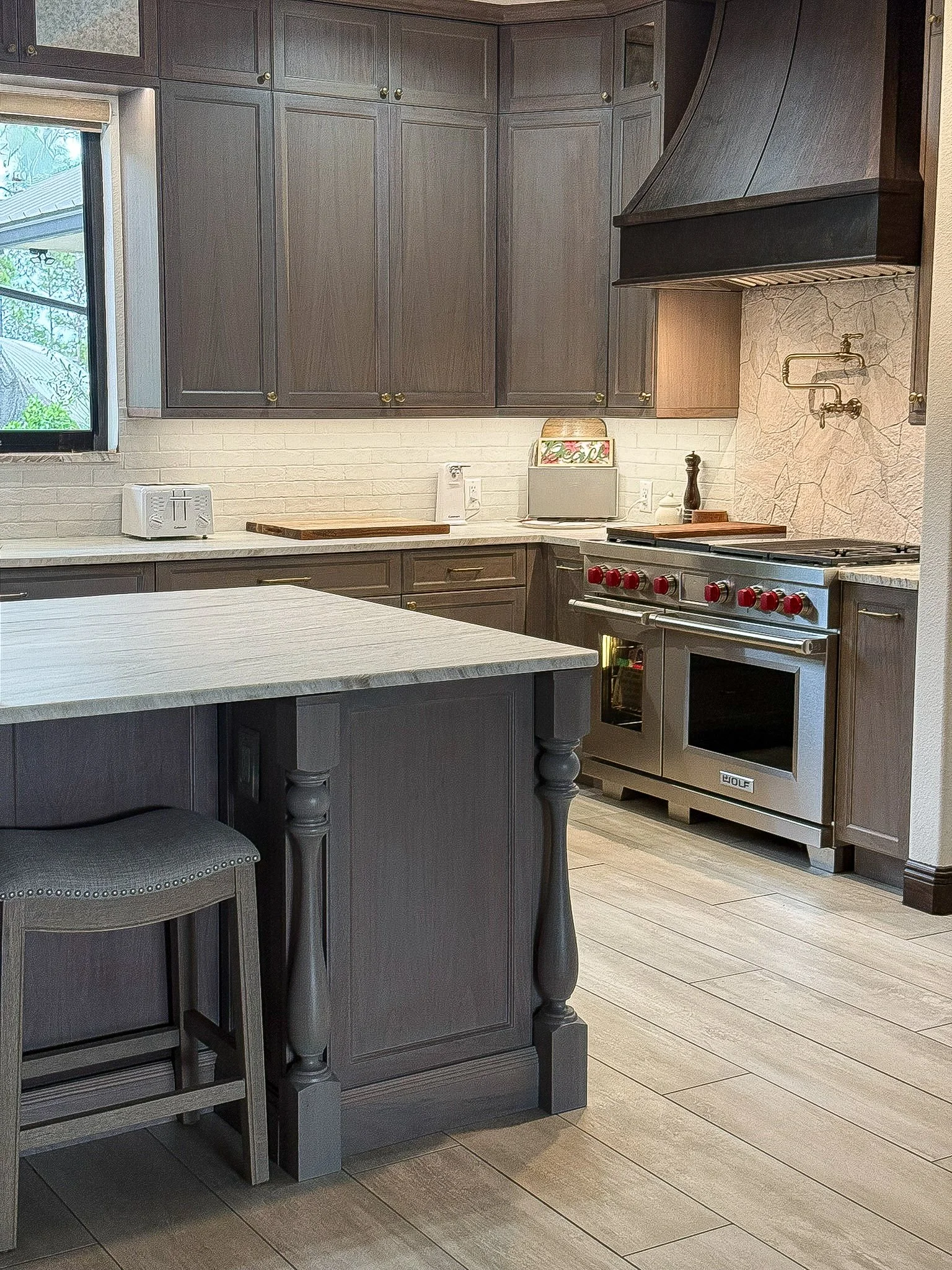 A cozy kitchen featuring dark gray cabinetry, a large island with a white countertop, a stainless steel Wolf oven with red knobs, a range hood, and a window with a view of greenery outside.