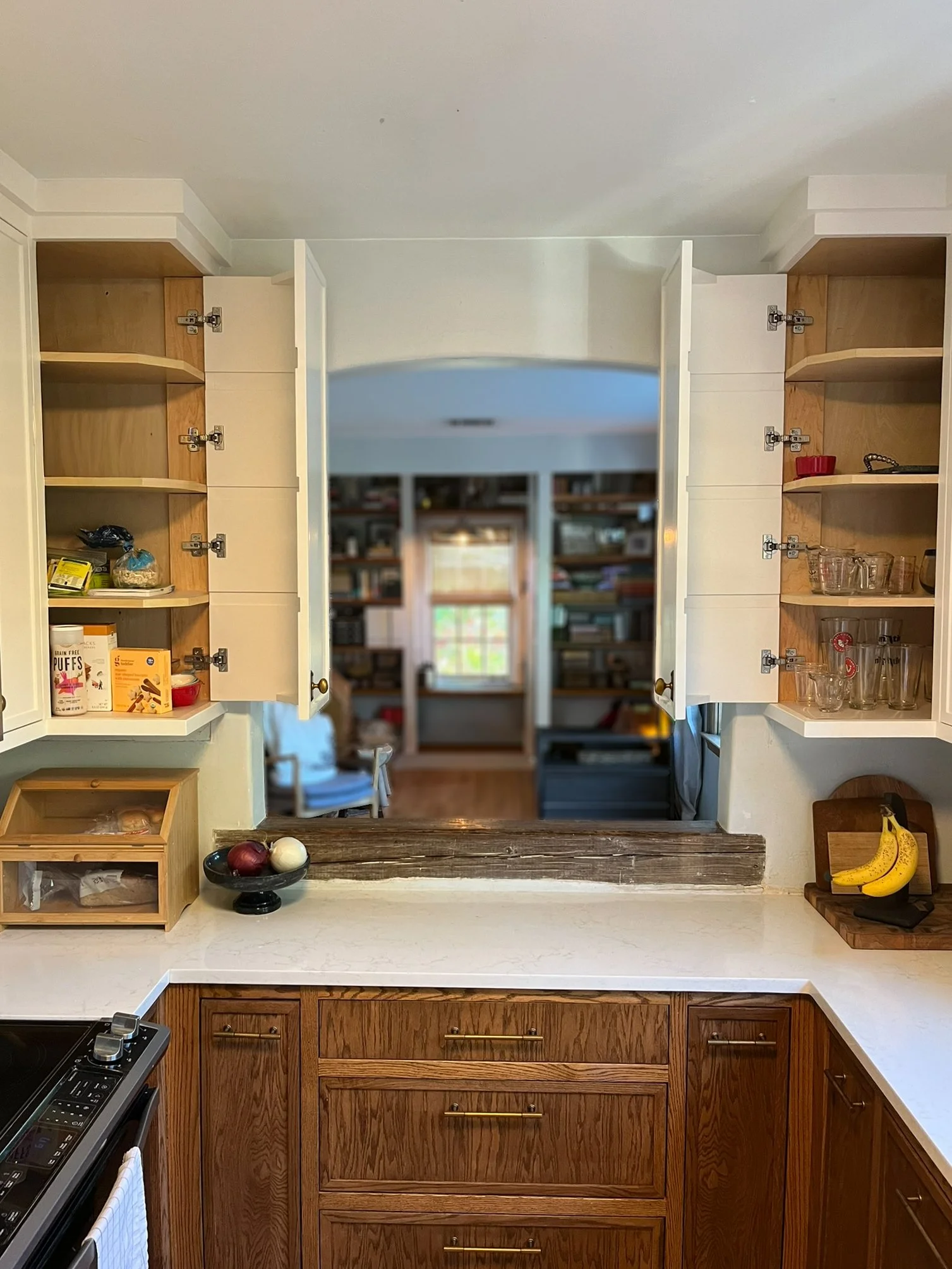 View of a kitchen with open upper cabinets, a cut-out pass-through window, and a view into a book-filled room. The countertop has a bowl of onions and a banana bunch, with a wooden box and a glass container nearby.