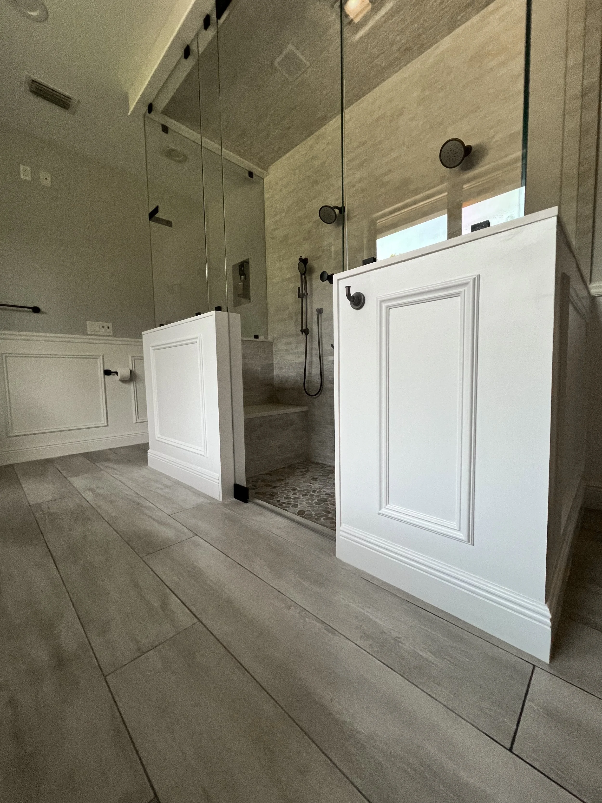 Modern walk-in shower with glass doors, black fixtures, and a pebble floor, in a bathroom with wood-look tile flooring and white wainscoting.