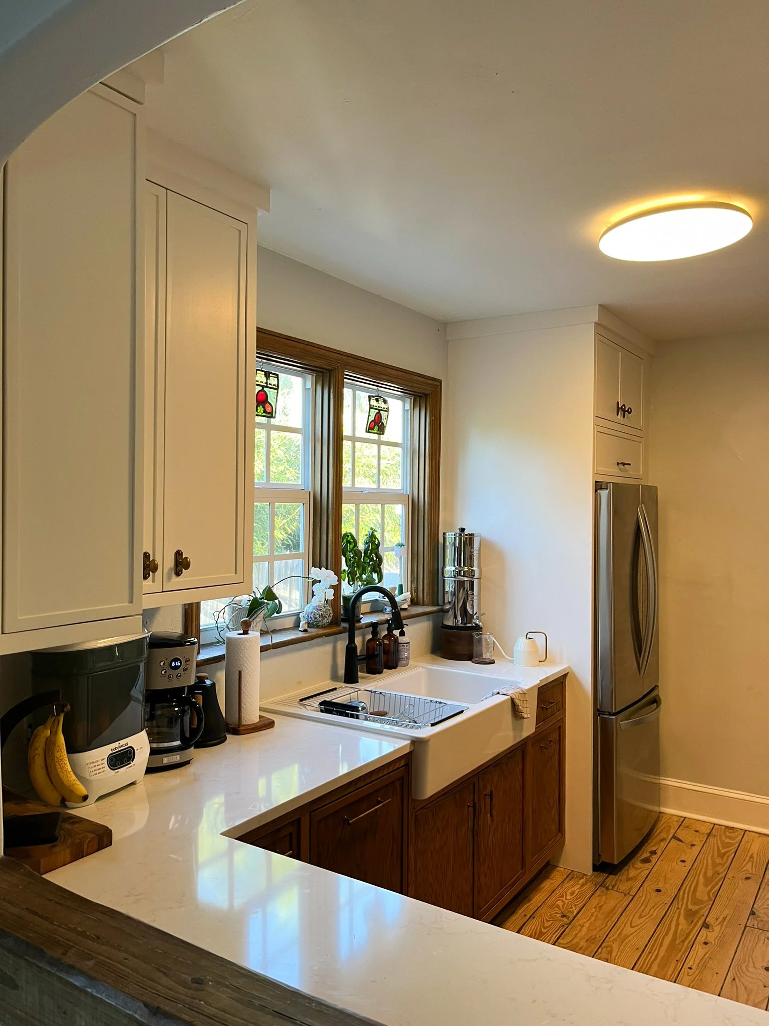 Kitchen with white upper cabinets, wood lower cabinets, double window, farmhouse sink, coffee maker, bananas, and stainless steel refrigerator.