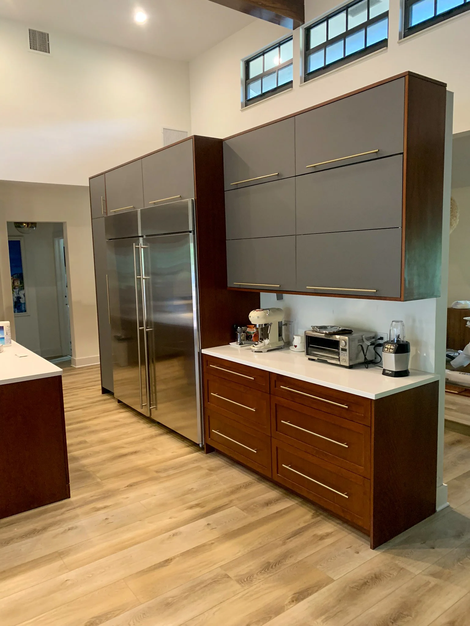 Modern kitchen with stainless steel refrigerator, dark upper cabinets, wooden lower cabinets, and small appliances on the white countertop.