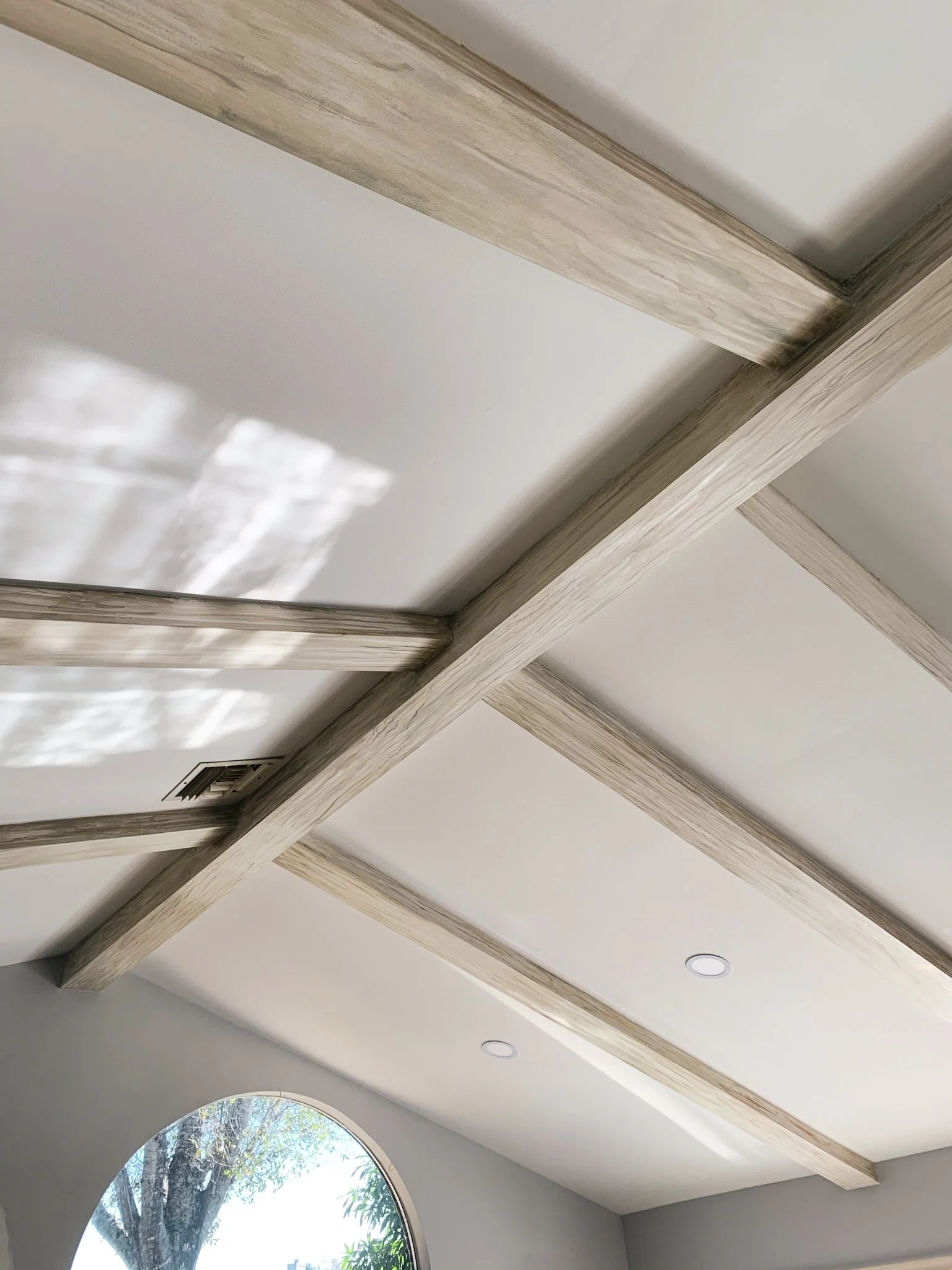 Ceiling with wooden beams, a corner with a circular window showing tree branches outside, and recessed lighting.