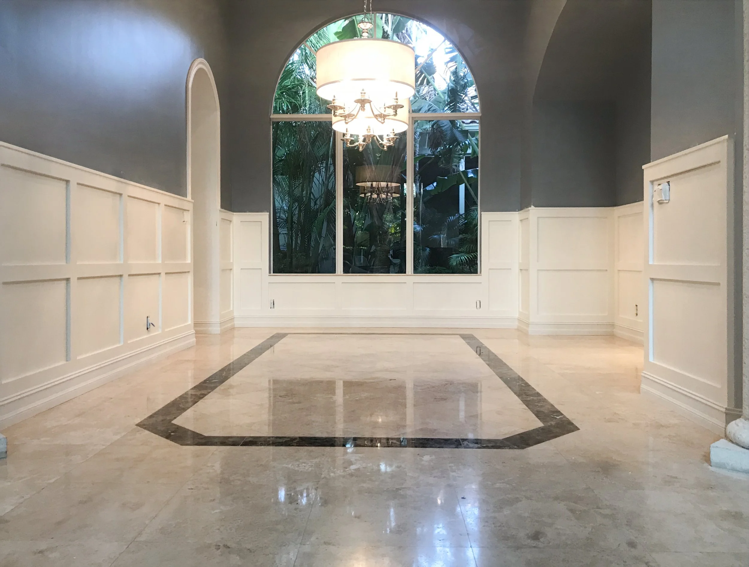 Empty elegant room with marble floor, wainscoting on walls, large window with greenery outside, and a chandelier hanging from the ceiling.