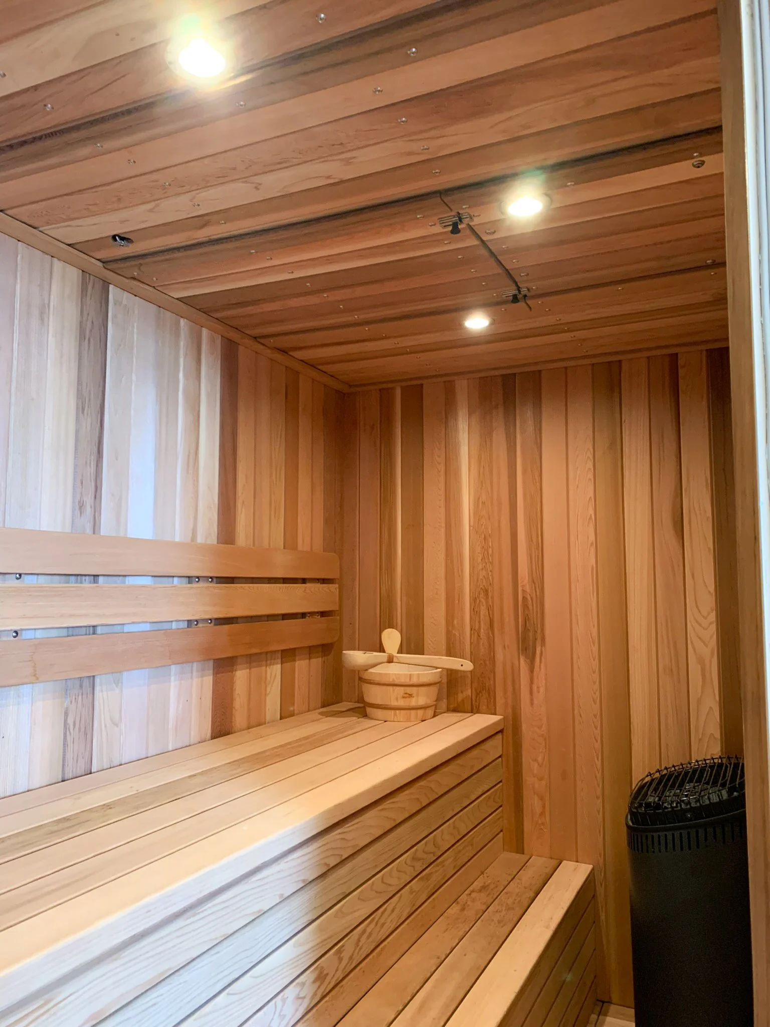 Interior of a wooden sauna with benches, a small wooden bucket with a ladle, and a black heater in the corner, illuminated by overhead lights.