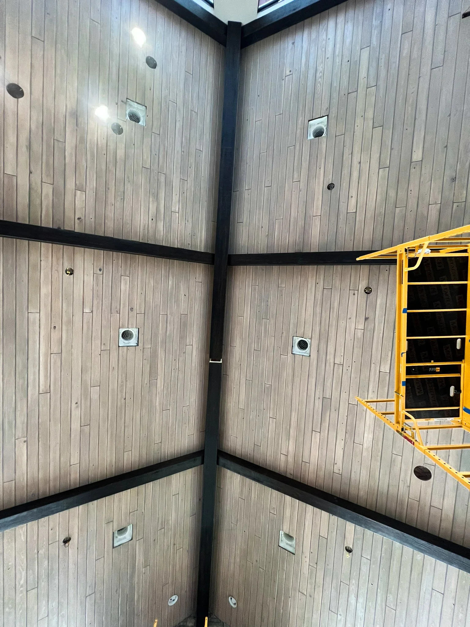 Ceiling of a gymnasium with black beams, wood paneling, and several exhaust vents. Part of a yellow scaffolding structure is visible on the right side.