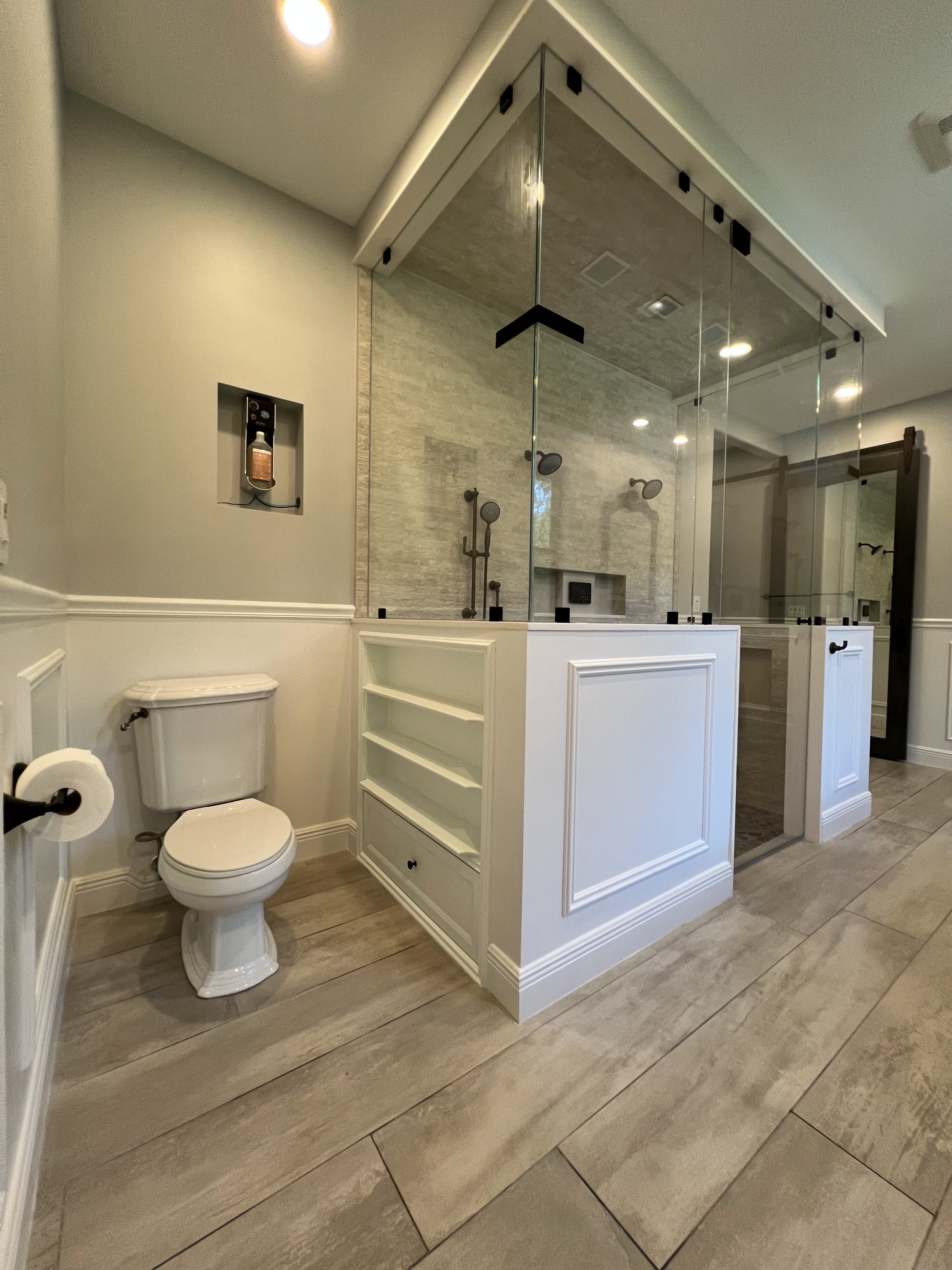Bathroom with a walk-in glass shower, toilet, decorative wall panels, and built-in shelves, with natural light and neutral colors.