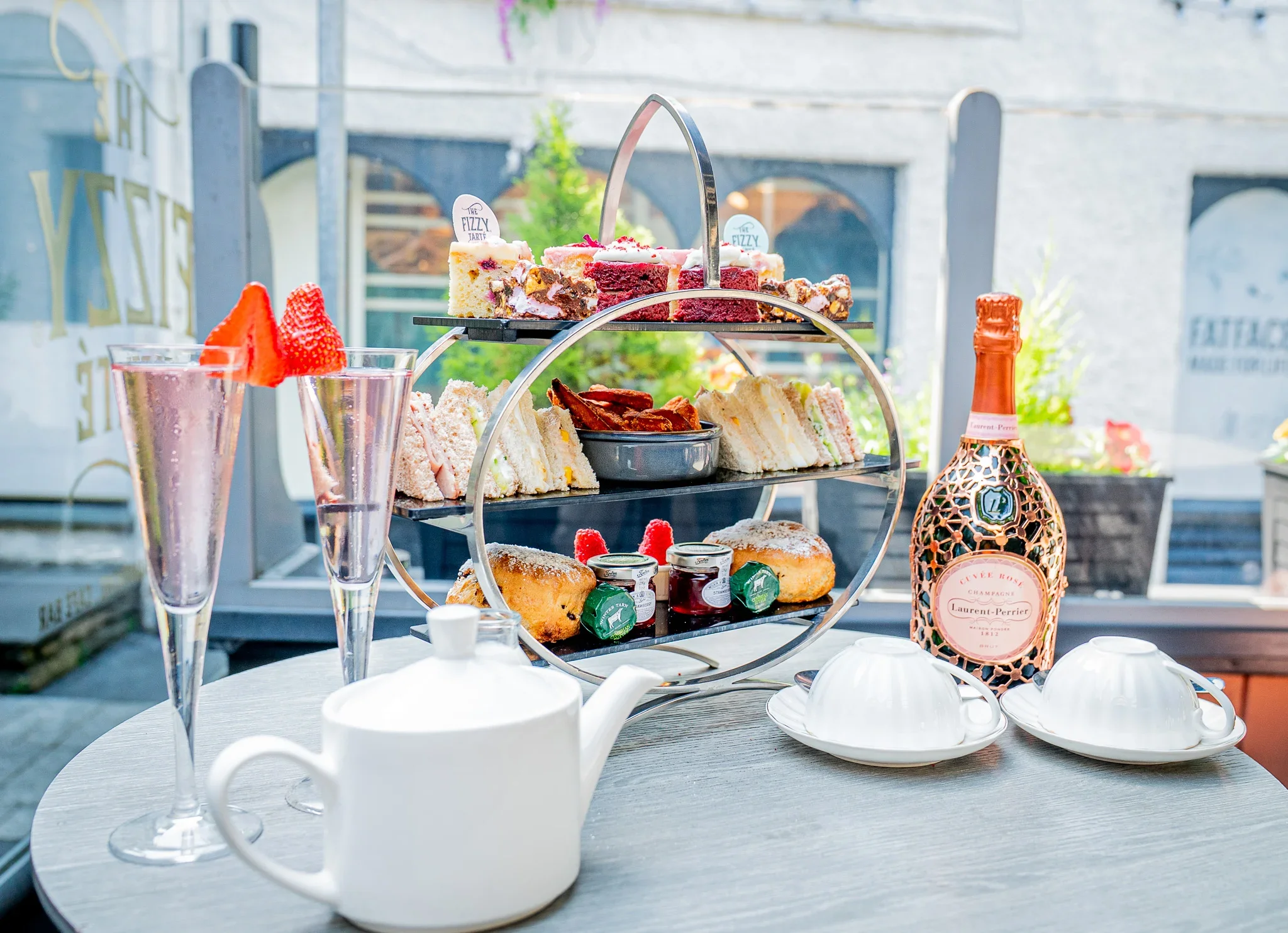 A complete three-tiered Laurent Perrier afternoon tea stand on a round table at The Fizzy Tarté in Lake district, featuring cakes, sandwiches, sweet potato fries, scones, and tea sets