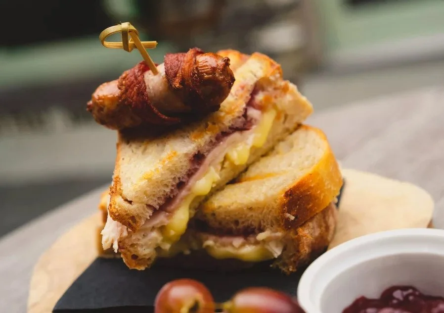 A close-up of a sandwich with bacon, melted cheese, and ham on toasted bread, held together with a toothpick, with a side of sauce and grapes in the background.