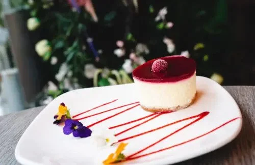 A round cheesecake with a red topping and a red raspberry on top, decorated with purple, yellow, and white edible flowers, and drizzled with red sauce on a white plate.
