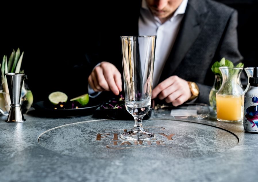 A bartender in a suit preparing a cocktail at a bar with various ingredients and tools, including a tall glass, lime, lemon juice, and herbs.