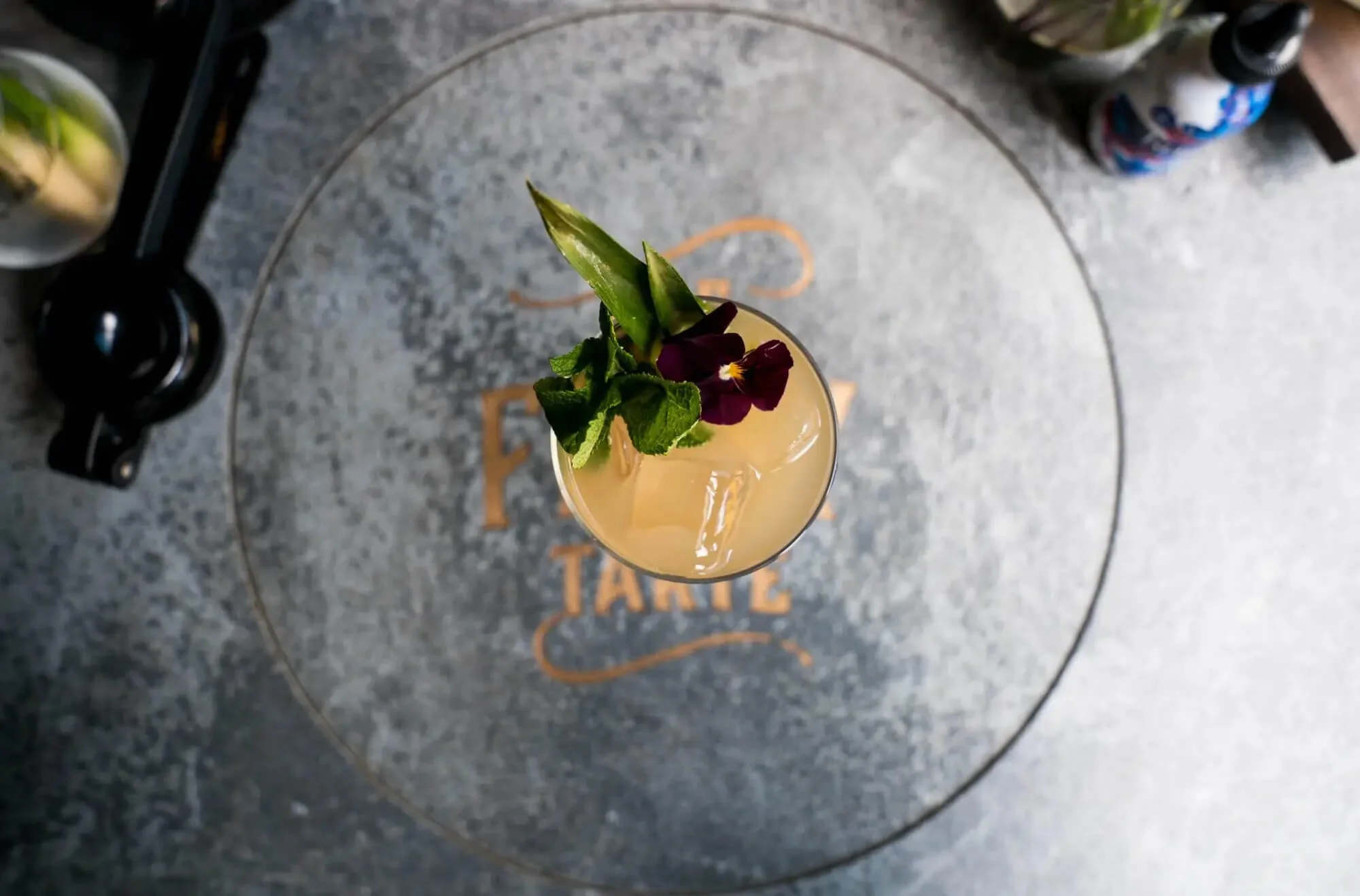 A tropical cocktail garnished with pineapple slices, a purple edible flower, and mint leaves, placed on a gray table with the words 'Funk Taco' written on it.