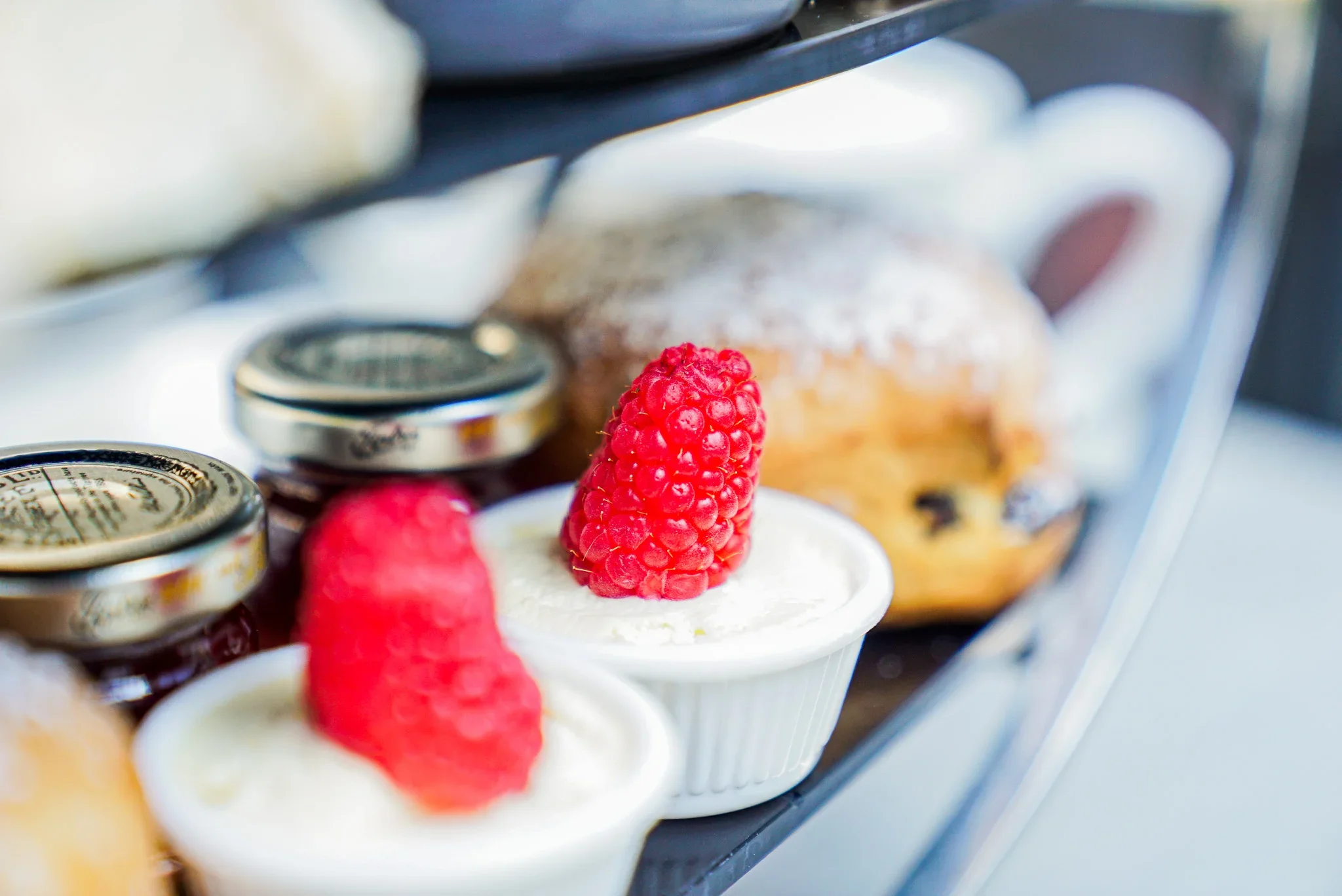 Close-up of fresh scones, miniature preserve jars, and ramekins of clotted cream topped with fresh raspberries on an afternoon tea tier at The Fizzy Tarté.
