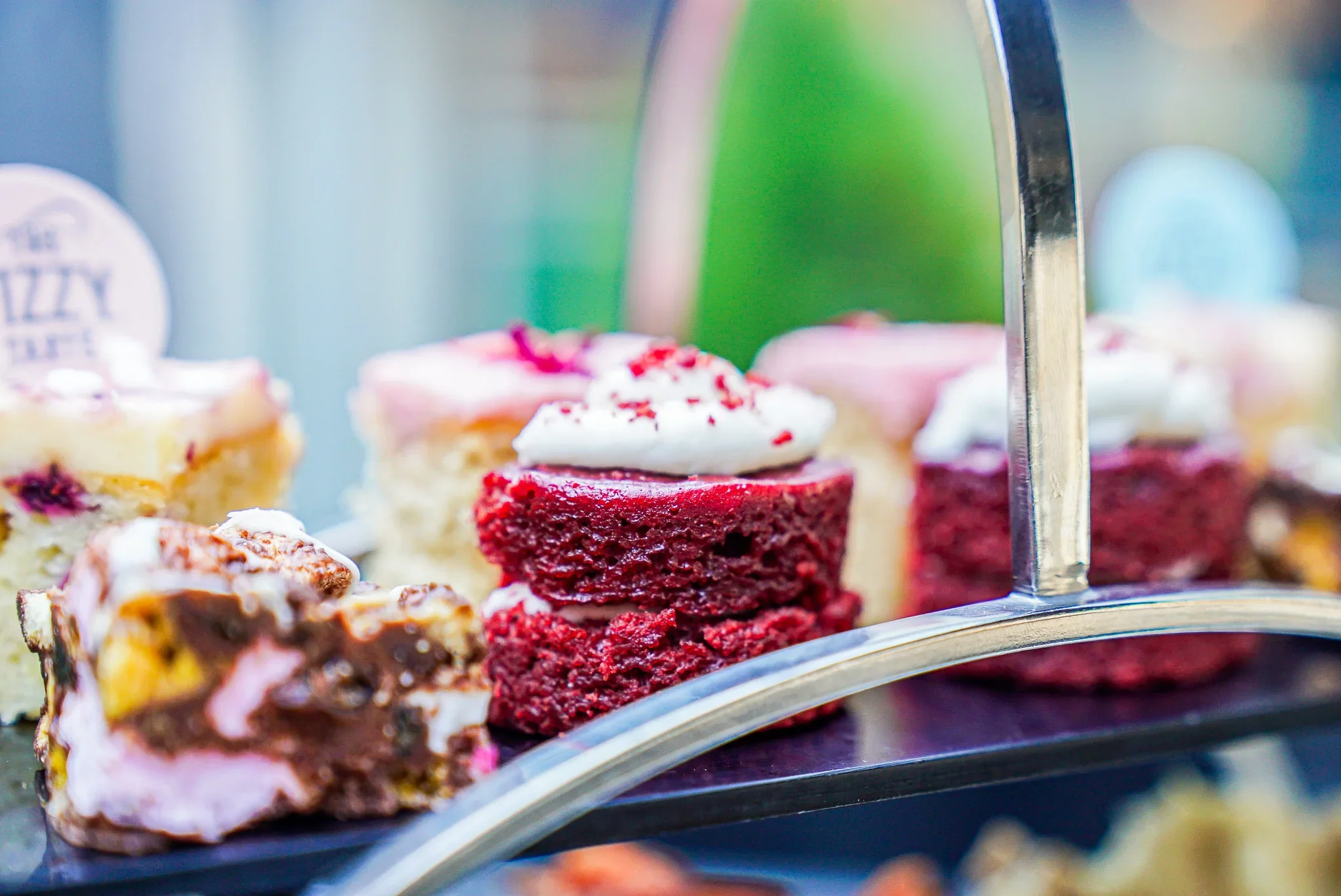 Close-up shot of a luxury afternoon tea tier at The Fizzy Tarté in Bowness-on-Windermere, featuring a mini red velvet cake with cream frosting, chocolate rocky road sweets, and pink glazed sponge cakes.