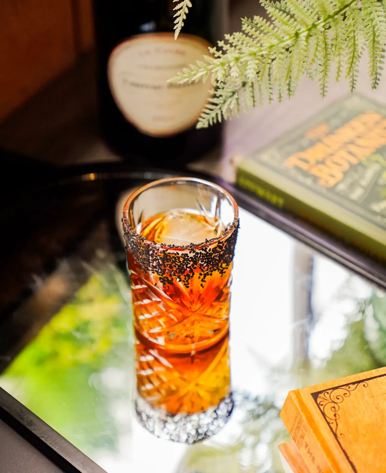 A tall glass with a patterned design and black sprinkles along the rim, filled with an orange-colored drink and ice, placed on a reflective surface near a window with greenery outside.