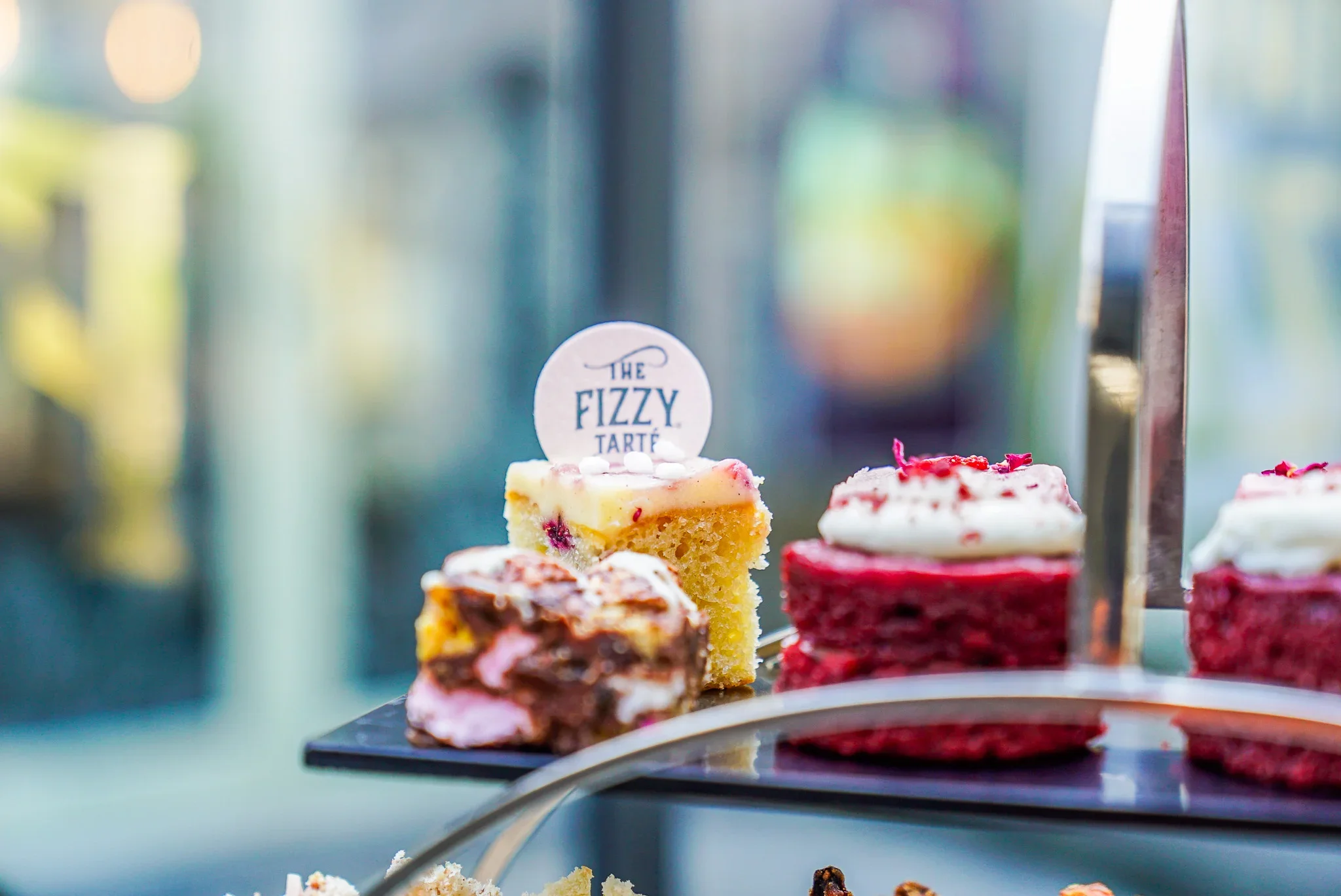 Close-up of top tier on an afternoon tea stand at The Fizzy Tarté in Bowness, showing a mini sponge cake topped with a white chocolate branded logo disc next to a red velvet cake.