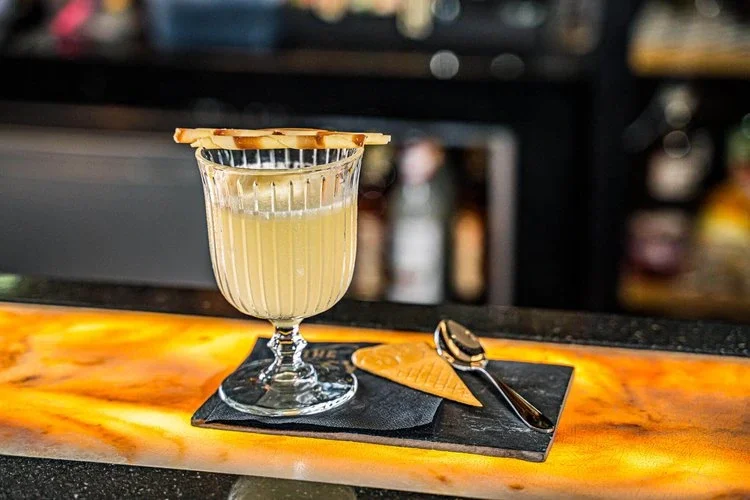 A glass of cocktail with a garnish, served on a black napkin, placed on a marble bar counter with a small spoon and a lemon wedge.